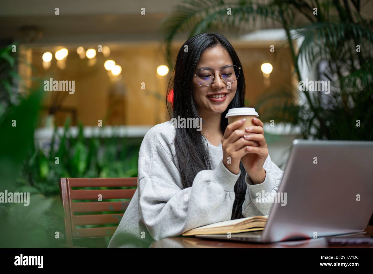 Una bella e rilassata donna asiatica in un intimo maglione si gode un caffè mentre lavora a distanza da una caffetteria. bevete, gente, stili di vita Foto Stock