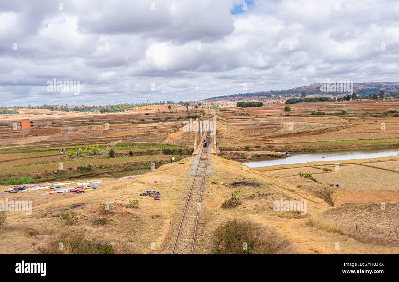 Ponte ferroviario panoramico ad arco che attraversa il fiume in praterie asciutte con un cielo nuvoloso drammatico. Paesaggio agricolo rurale con campi terrazzati e sparsi Foto Stock