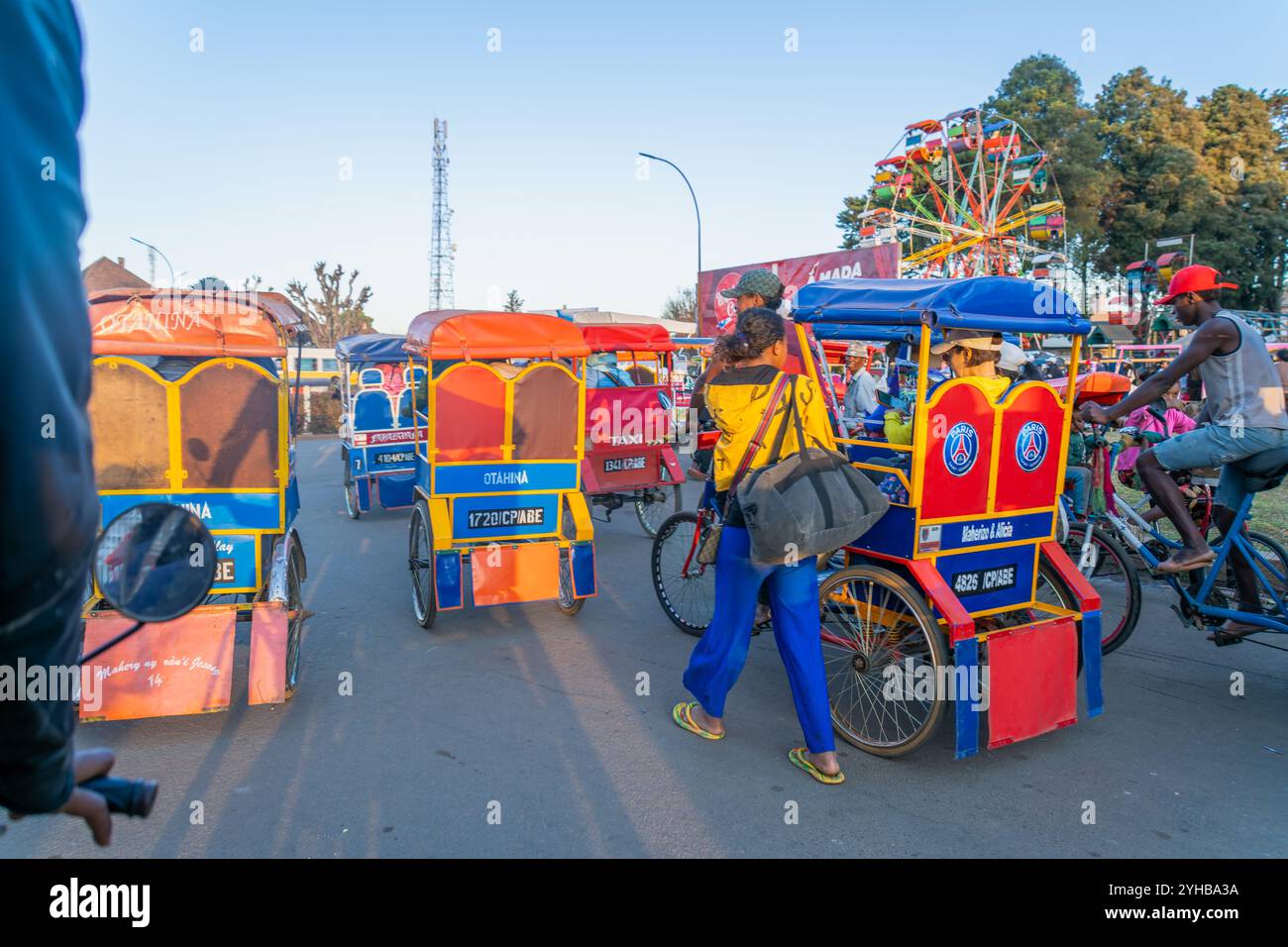 Antsirabe, Madagascar - 30 agosto 2024: Colorati risciò tradizionali si allineano vicino alla ruota panoramica in questa città di altipiani. Questi risciò sono un pop Foto Stock