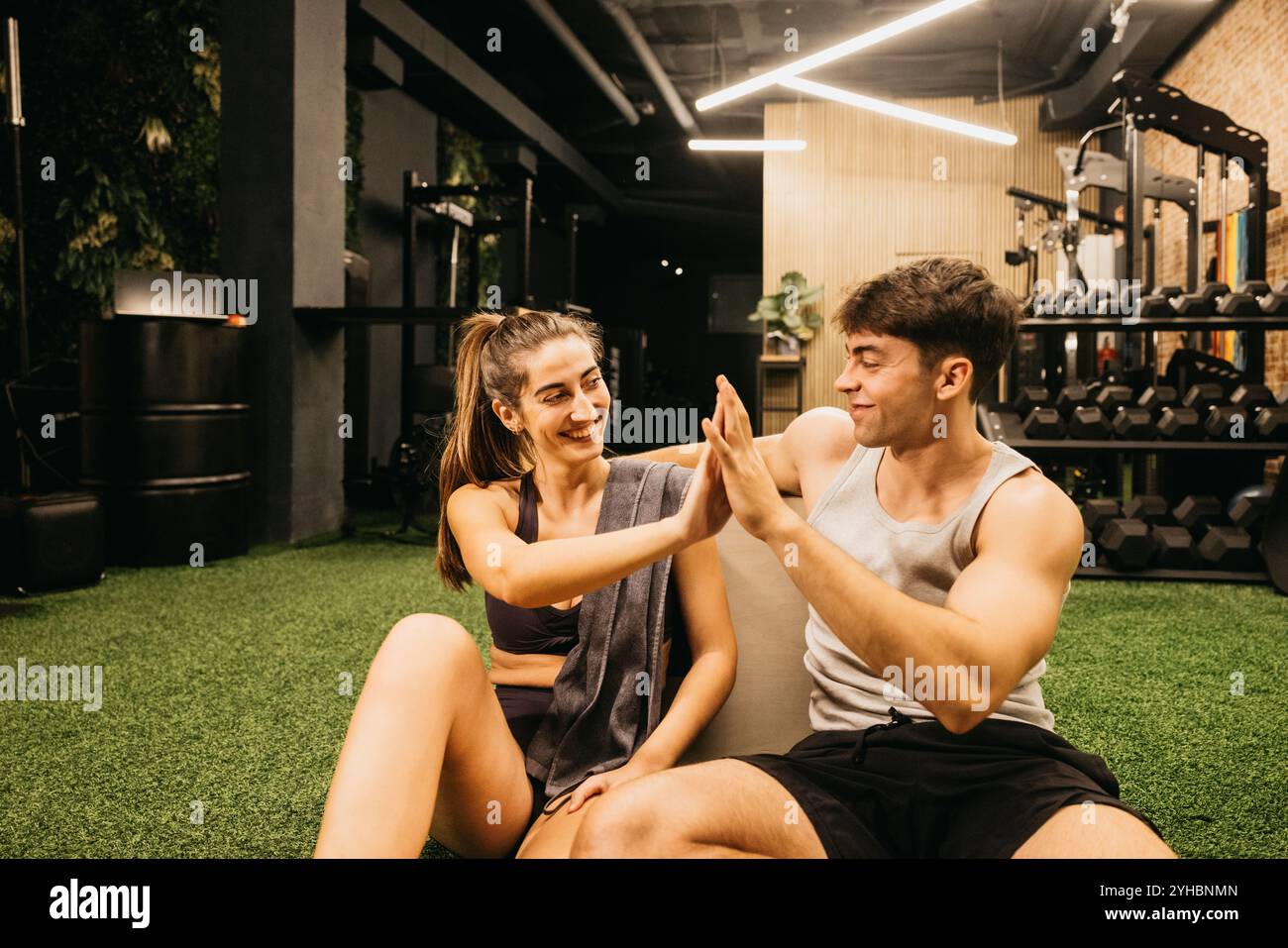 Una coppia allegra si siede sul pavimento della palestra, condividendo cinque alti dopo un allenamento. L'ambiente e' una moderna palestra con attrezzature per l'esercizio fisico sullo sfondo. Foto Stock