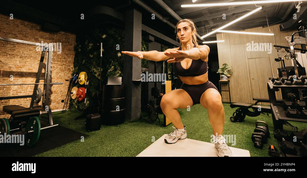 Donna che esegue squat in un ambiente moderno con attrezzature visibili. La sua espressione mostra dedizione e concentrazione, sottolineando un impegno Foto Stock