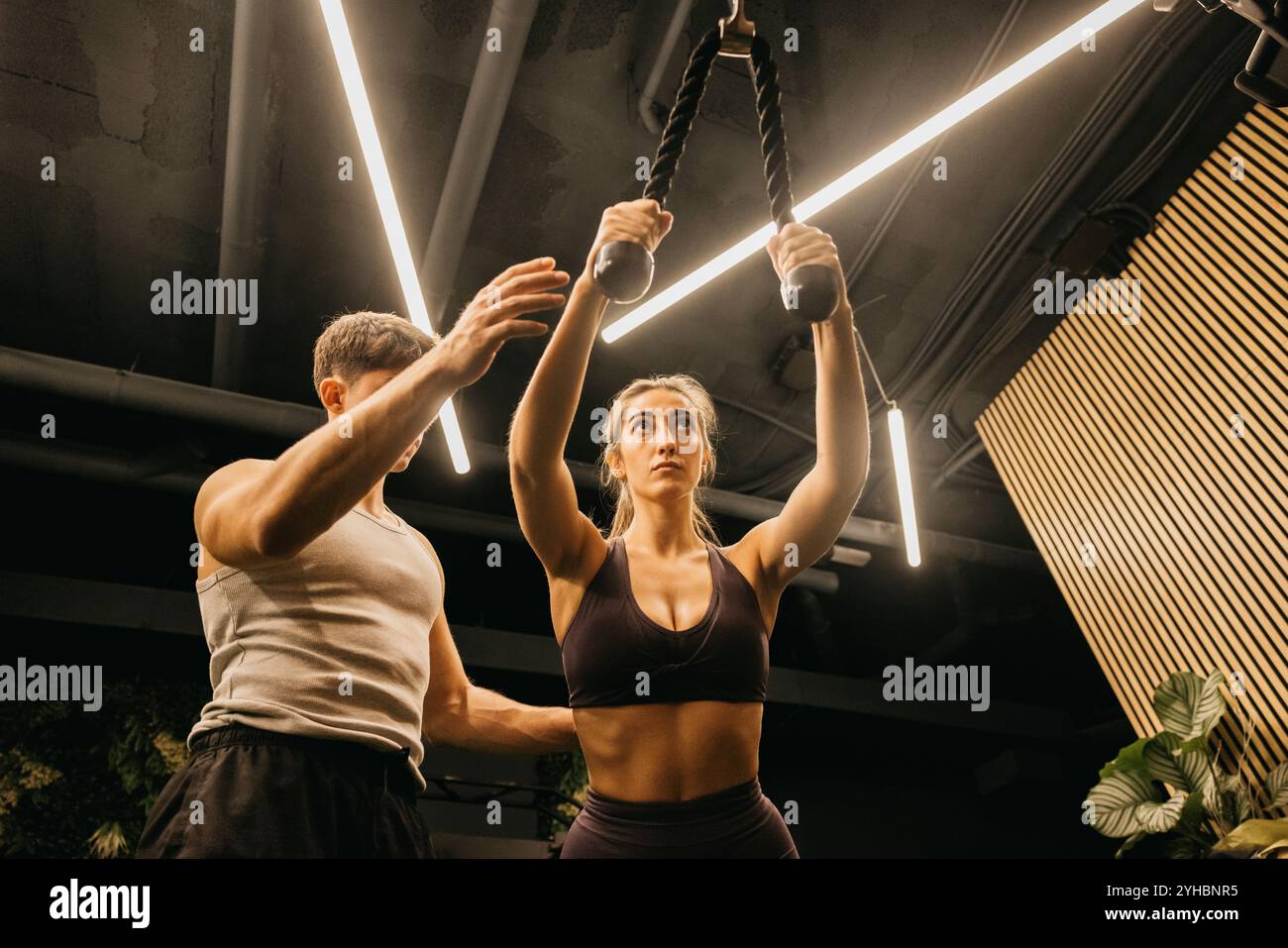 Una donna concentrata si allena con un allenatore di fitness in palestra, usando esercizi di corda per la forza della parte superiore del corpo. Foto Stock