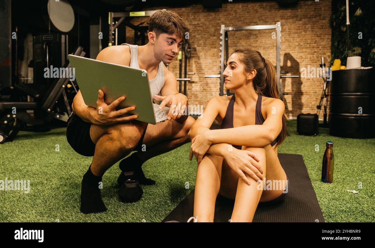 Il formatore e il cliente discutono della strategia di fitness utilizzando un computer portatile in palestra, promuovendo la motivazione e la pianificazione degli allenamenti. Foto Stock