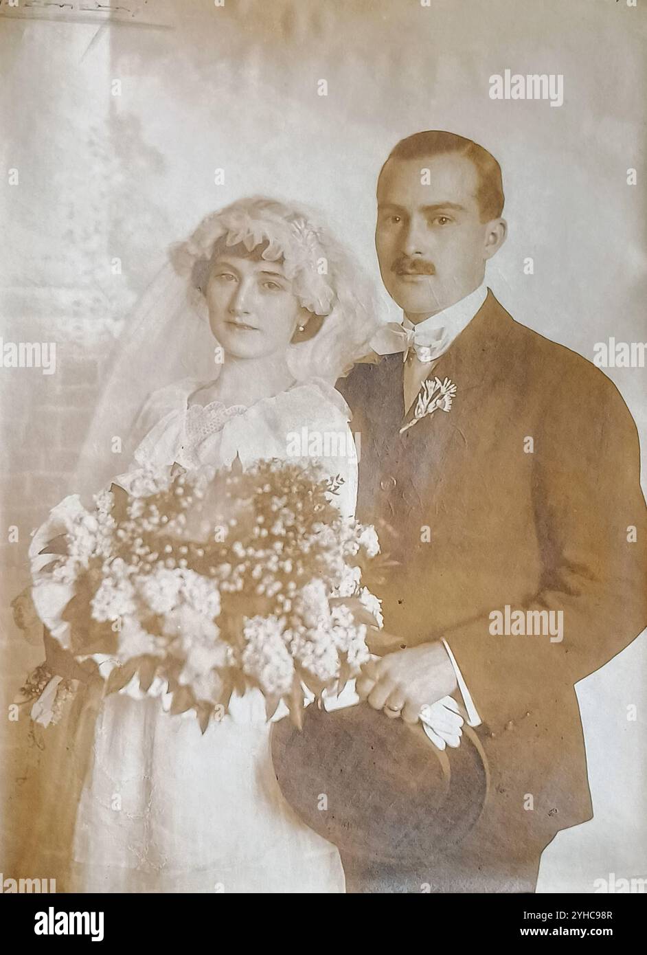 Jugoslavia - 1919: Ritratto di nozze di una bella coppia di sposi novelli in piacevoli tonalità seppia; sposa in abito da sposa bianco, con velo bianco che regge un grande bouquet di fiori bianchi, sposo con un baffo in piedi dietro di lei indossa un abito scuro con un fiore nel suo risvolto, cravatta con fiocco bianco e tenendo in mano un cappello da bowler e guanti da whote Foto Stock