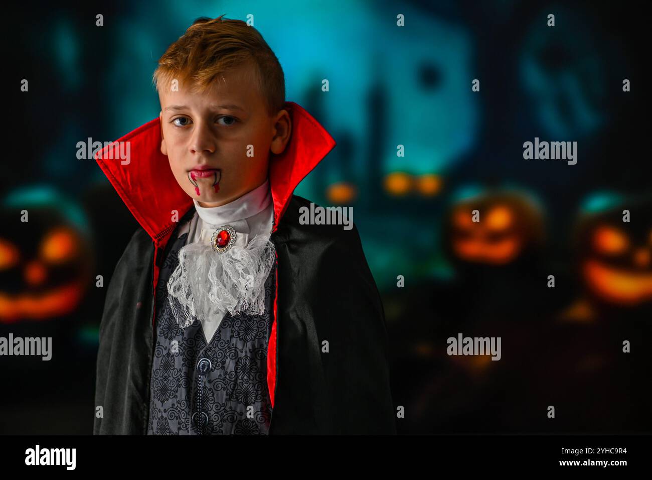 Bambino vestito da vampiro che posa di fronte a uno sfondo sfocato con zucche e alberi scuri. Foto Stock