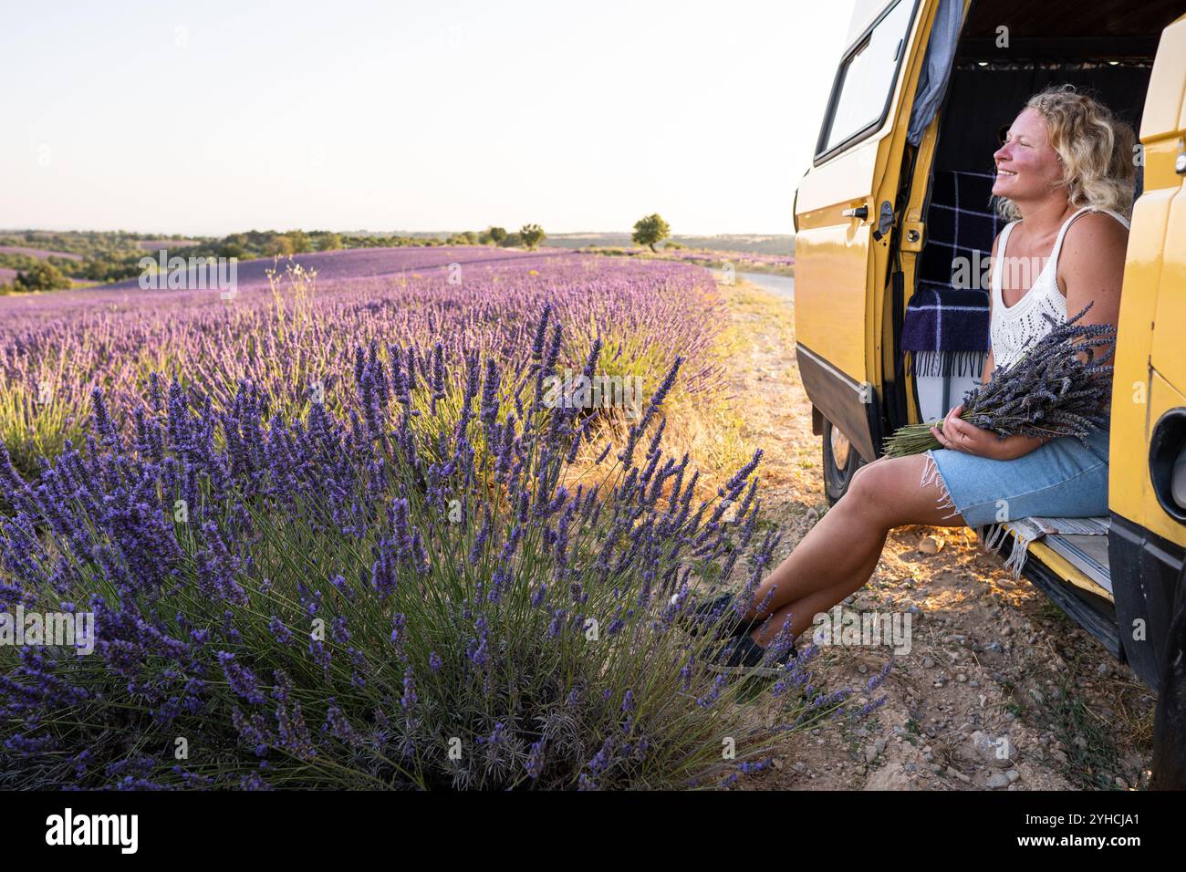 Giovane donna bionda che si gode la vista sul suo camper giallo rétro all'alba nei campi di lavanda di Valensole in Provenza, nel sud della Francia Foto Stock