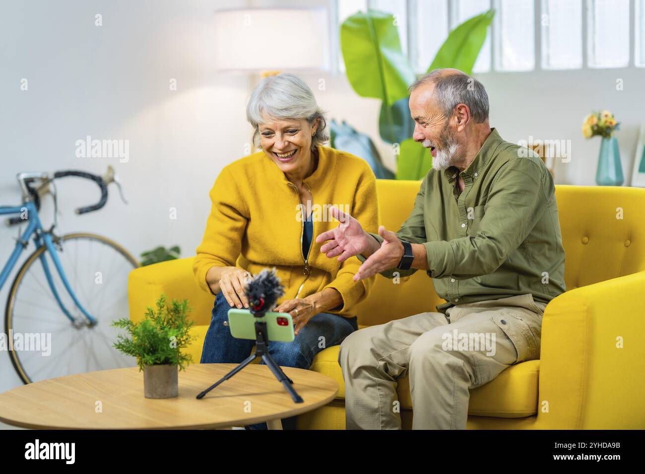 Coppia senior sorridente durante una videochiamata utilizzando lo smartphone seduto su un moderno divano giallo di casa Foto Stock