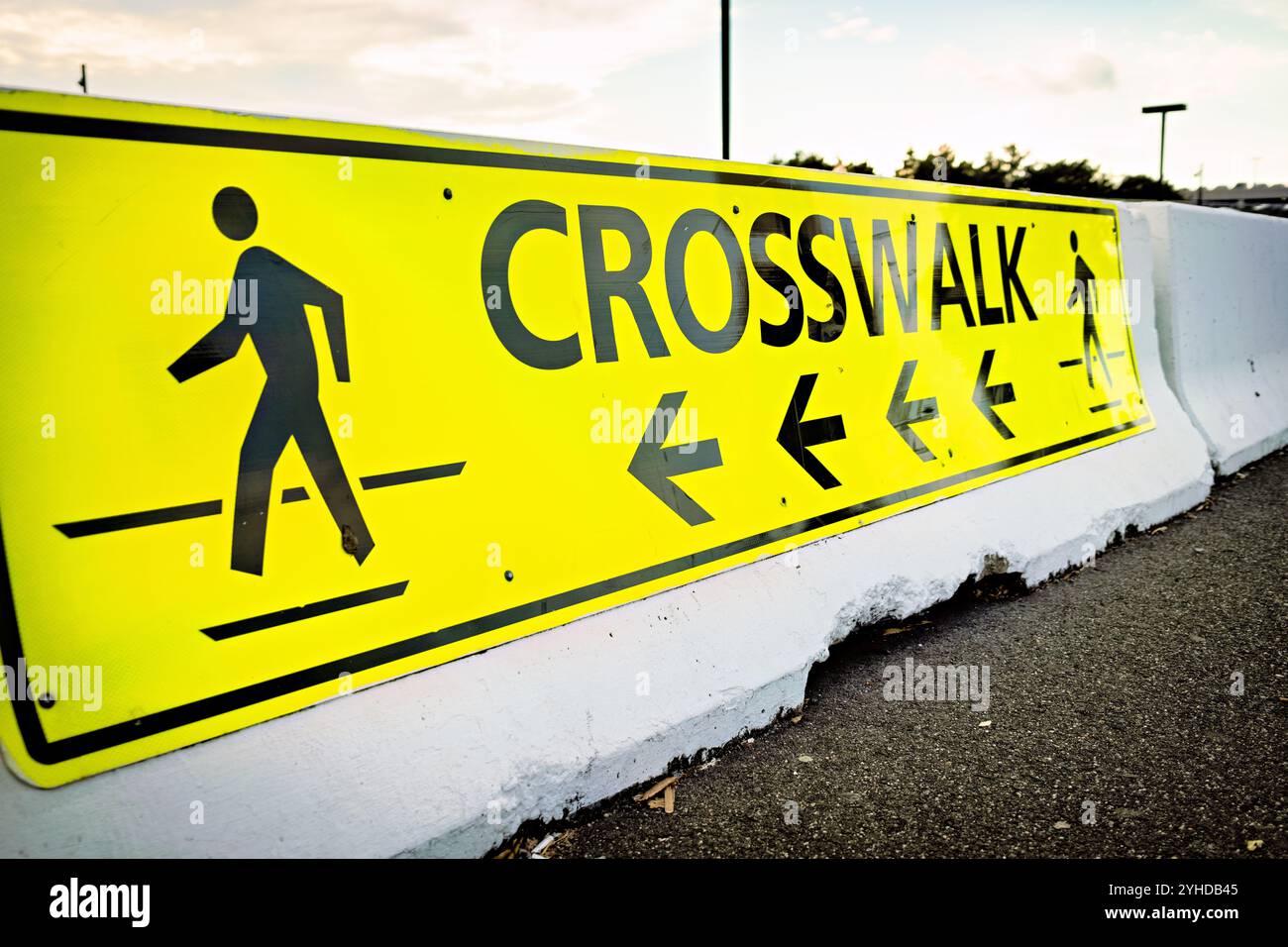 Segnale di attraversamento pedonale Dulles Virginia // DULLES, Virginia - Un segnale giallo luminoso di attraversamento pedonale montato su una barriera in cemento fornisce avvertenze ad alta visibilità ai conducenti e ai pedoni presso una struttura di trasporto. L'attrezzatura per la sicurezza stradale è dotata del simbolo di pedone standard su un segnale di avvertenza a forma di diamante, fissato a una barriera stradale in cemento. Questo tipo di infrastruttura di sicurezza è comunemente utilizzato in tutti i nodi di trasporto per dirigere il traffico pedonale e migliorare la sicurezza dei pedoni nelle aree in cui il veicolo è in movimento. La colorazione gialla ad alto contrasto è progettata per offrire un massimo di Foto Stock