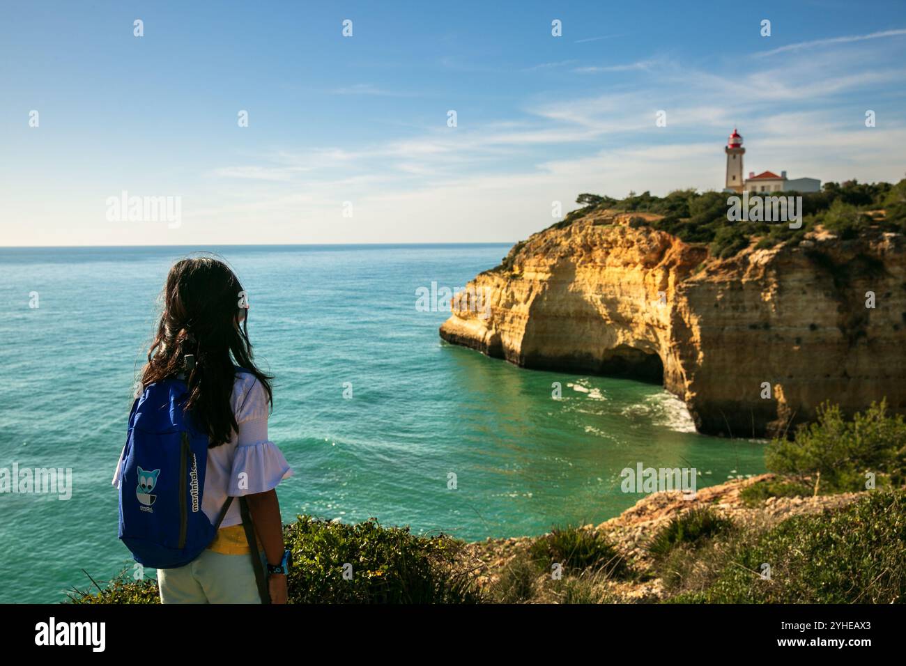 Una giovane ragazza ammira il faro di Alfanzina da un sentiero escursionistico in una giornata di sole, incorniciato dalle aspre scogliere e dall'oceano turchese dell'Algarve Foto Stock