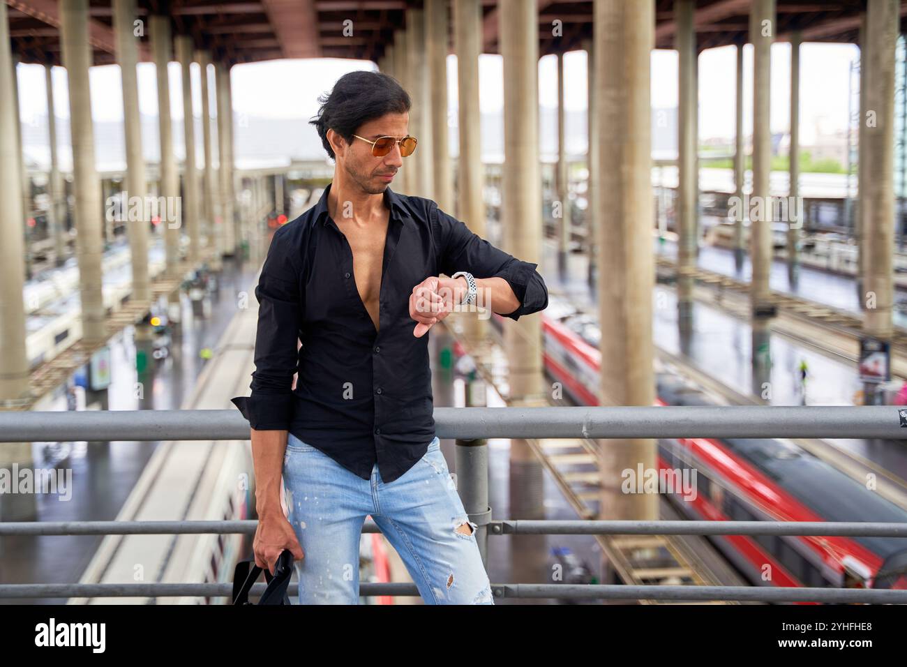 Uomo indiano con una camicia nera e jeans strappati controllando il suo orologio in una stazione ferroviaria, tenendo gli occhiali da sole, con treni e piattaforme sullo sfondo. Foto Stock