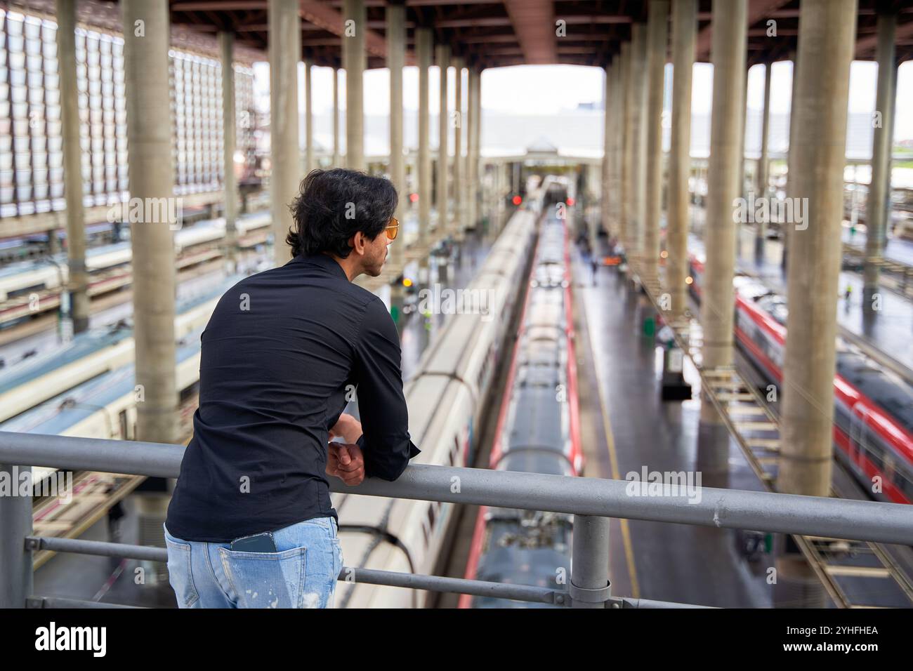 Uomo indiano in camicia nera e jeans strappati che guarda da un ponte la piattaforma di un treno. Foto Stock