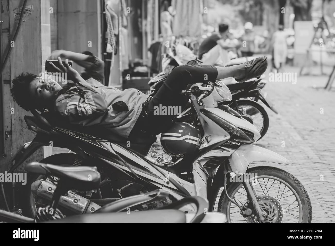 Un giovane si rilassa casualmente su una moto, scorrendo sul telefono, catturando un momento di pace nella trafficata strada di Hanoi, in bianco e nero. Foto Stock