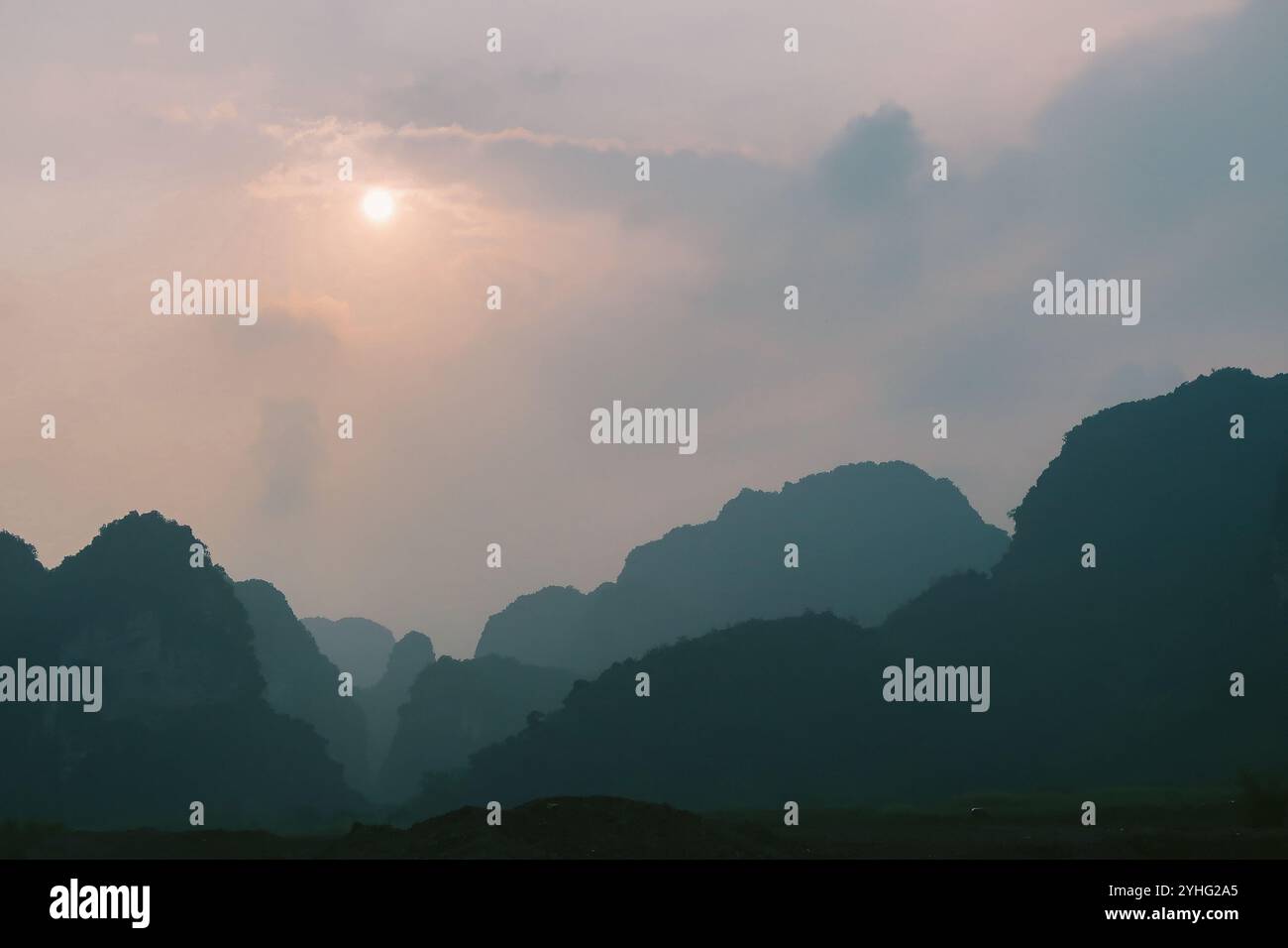 Il sole nebbioso sorge sulle montagne carsiche di Ninh Binh, Vietnam, che proietta un bagliore tranquillo attraverso le morbide nuvole e crea un paesaggio sereno e suggestivo Foto Stock