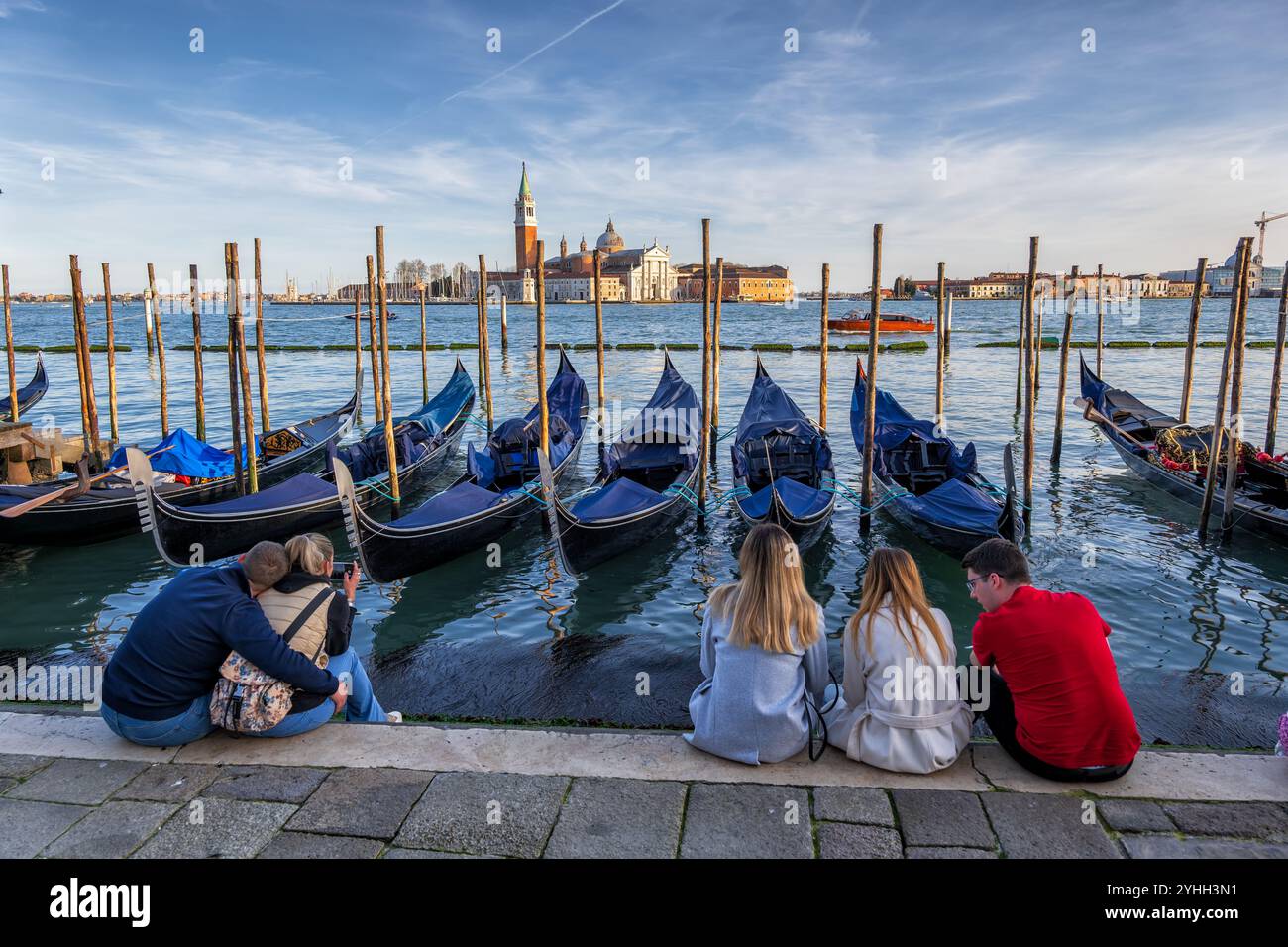 Città di Venezia, Italia, persone sedute sul lungomare di Riva degli Schiavoni, che guardano le iconiche gondole e le tradizionali barche e l'isola di San Giorgio maggiore Foto Stock