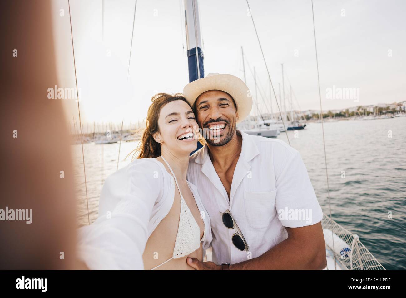 Una coppia sorridente scatta un selfie mentre si rilassa su una barca a vela, godendosi una giornata di sole al porto. Perfetto momento di vacanza estiva per catturare gioia e amore. Foto Stock