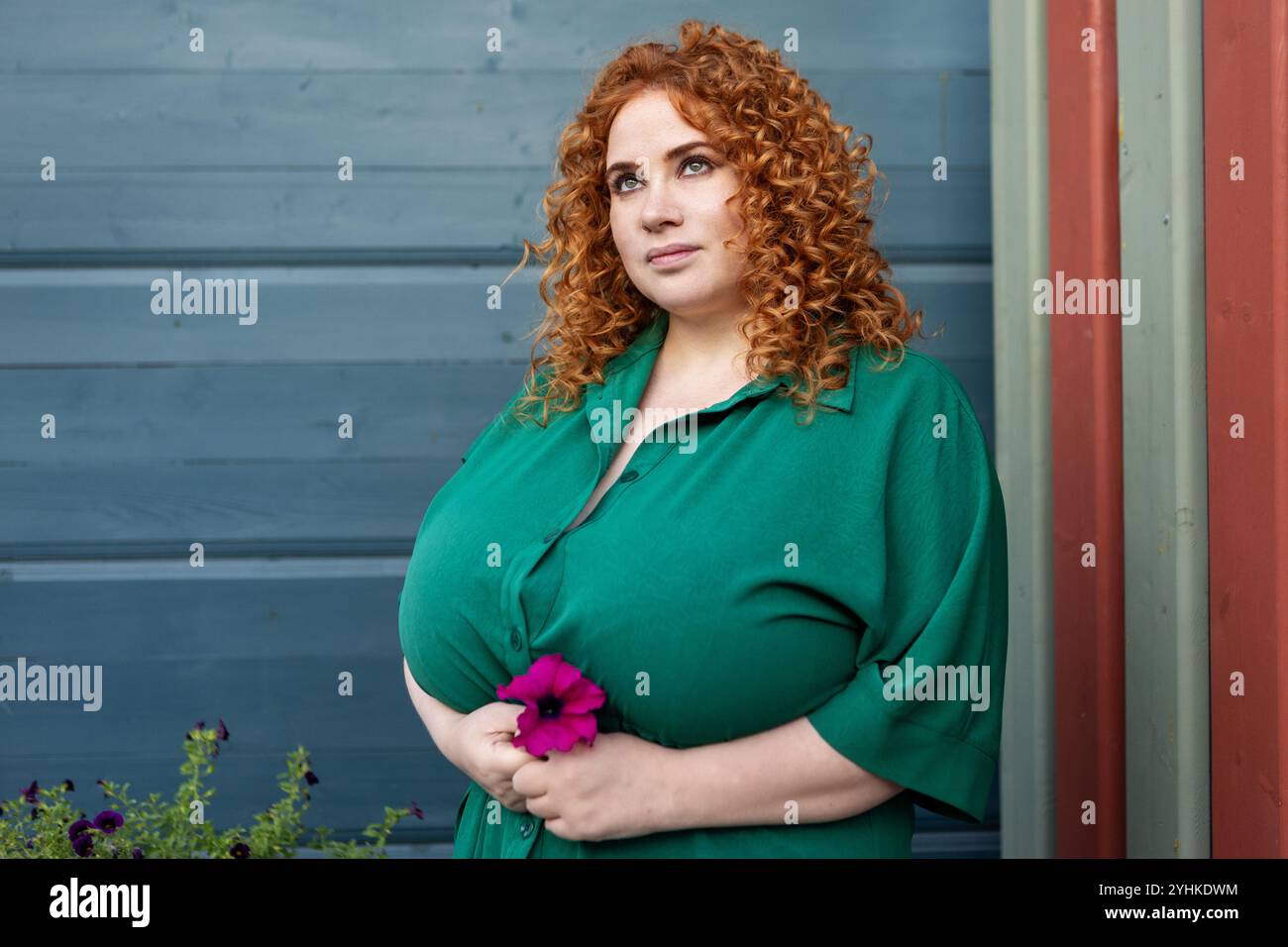 Plus size Woman con capelli ricci rossi in abbigliamento verde mantiene il fiore sullo sfondo blu della vivace parete, bellezza e sicurezza all'aperto Foto Stock