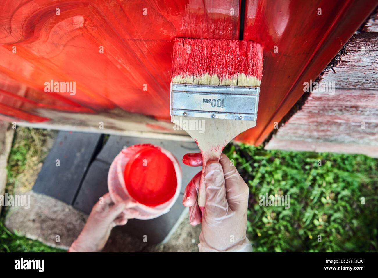 il pennello piatto largo 100 mm viene utilizzato per verniciare la porta d'ingresso in acciaio di una casa di campagna in legno di colore rosso. Foto Stock