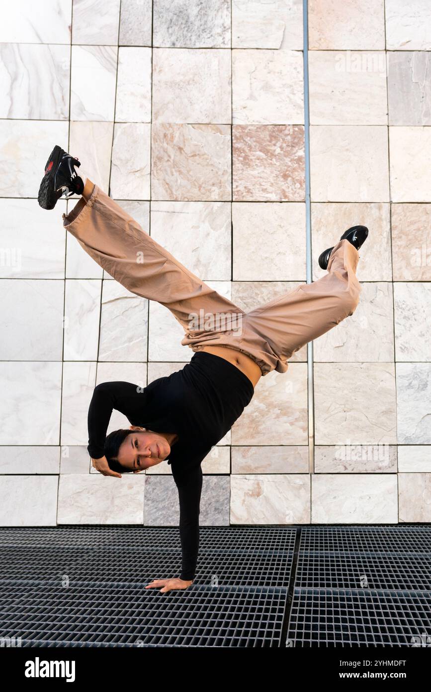 Una giovane donna con pantaloni abbronzati e t-shirt nera breakdance che esegue un cavalletto su una griglia di metallo. Concetto di atletismo e abilità, come la donna è ab Foto Stock