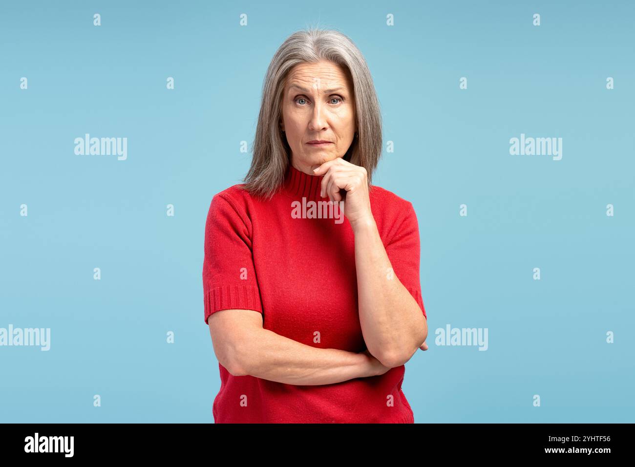 Donna dai capelli grigi, matura e seria, con un maglione rosso che pensa, guarda la fotocamera, posa isolata su sfondo blu. Concetto di pubblicità Foto Stock