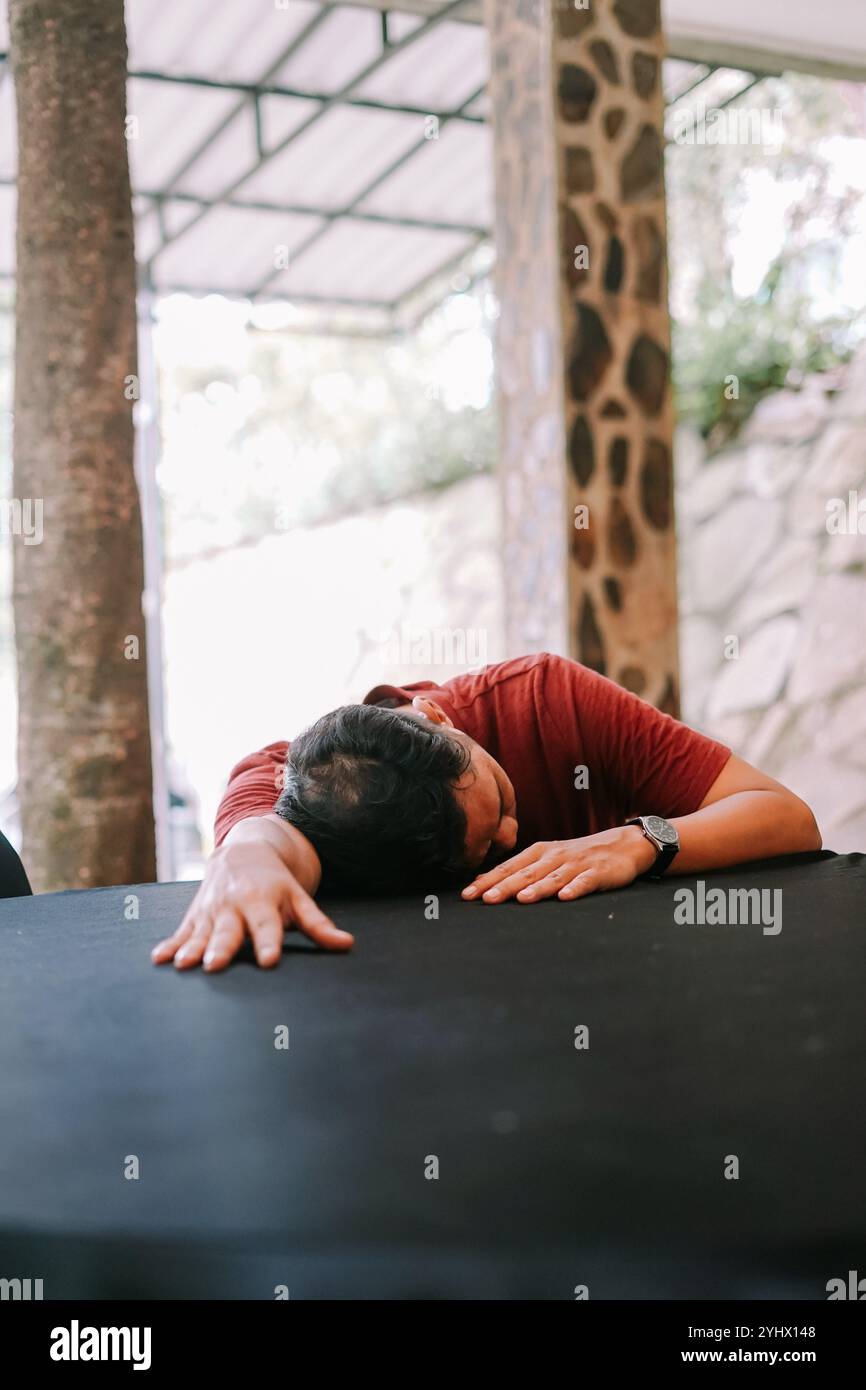 Un uomo con una t-shirt rossa si appoggia in avanti su un tavolo nero con la testa appoggiata e le braccia tese. La posa trasmette sfinimento, rilassamento o c Foto Stock