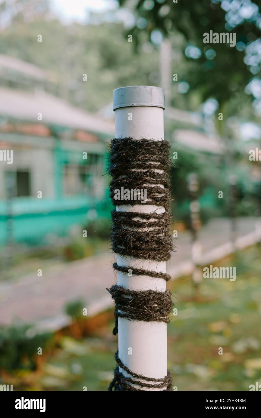 un palo bianco avvolto da una corda nera ruvida che crea un look testurizzato. Lo sfondo e' sfocato, caratterizzato da vegetazione e da un edificio, che dona un naturale, Foto Stock