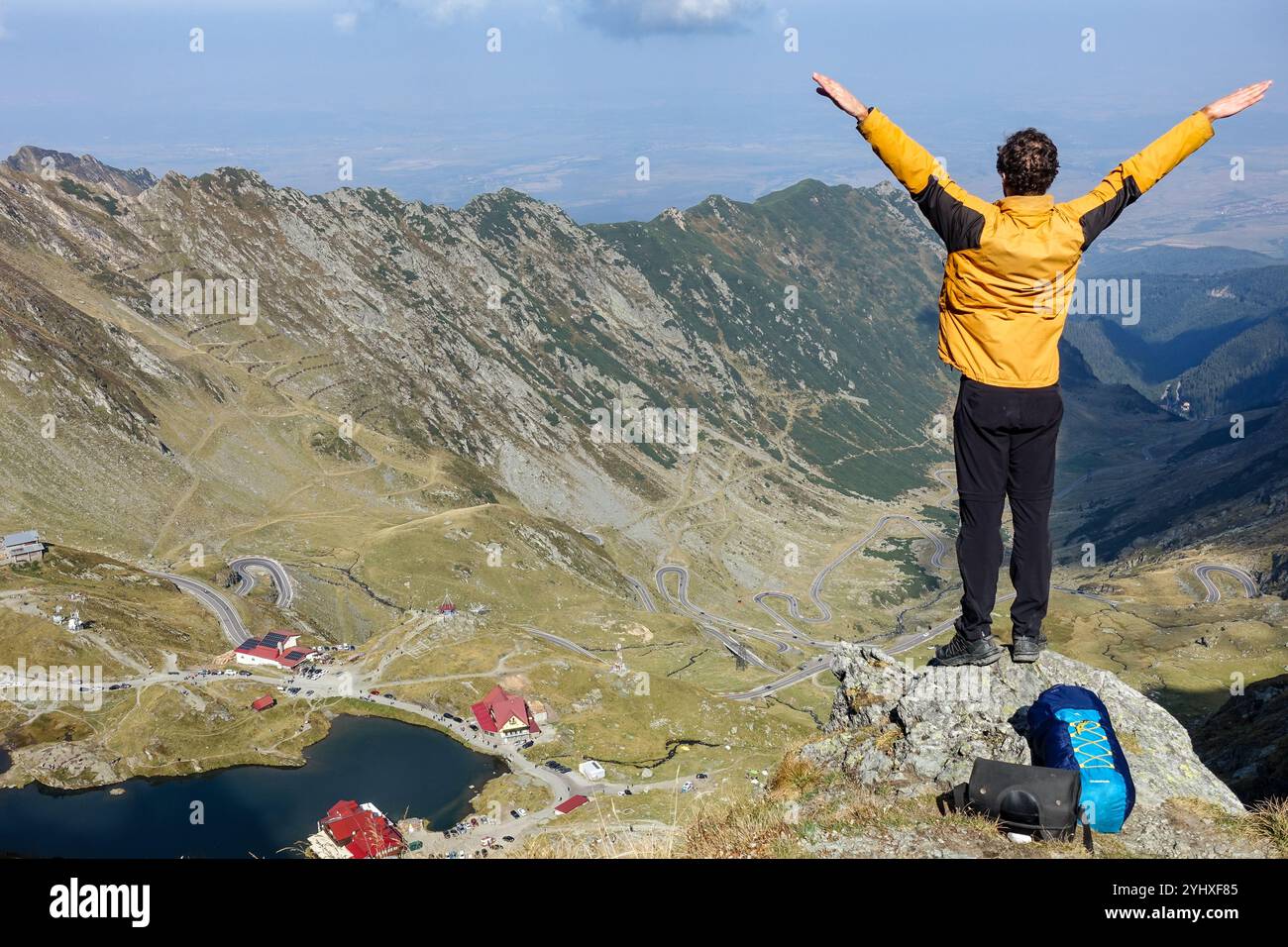 Escursionista che si erge trionfalmente sulla vetta della montagna che si affaccia sulla tortuosa strada Transfagarasan e sulla valle alpina vicino al lago Balea, Romania Foto Stock