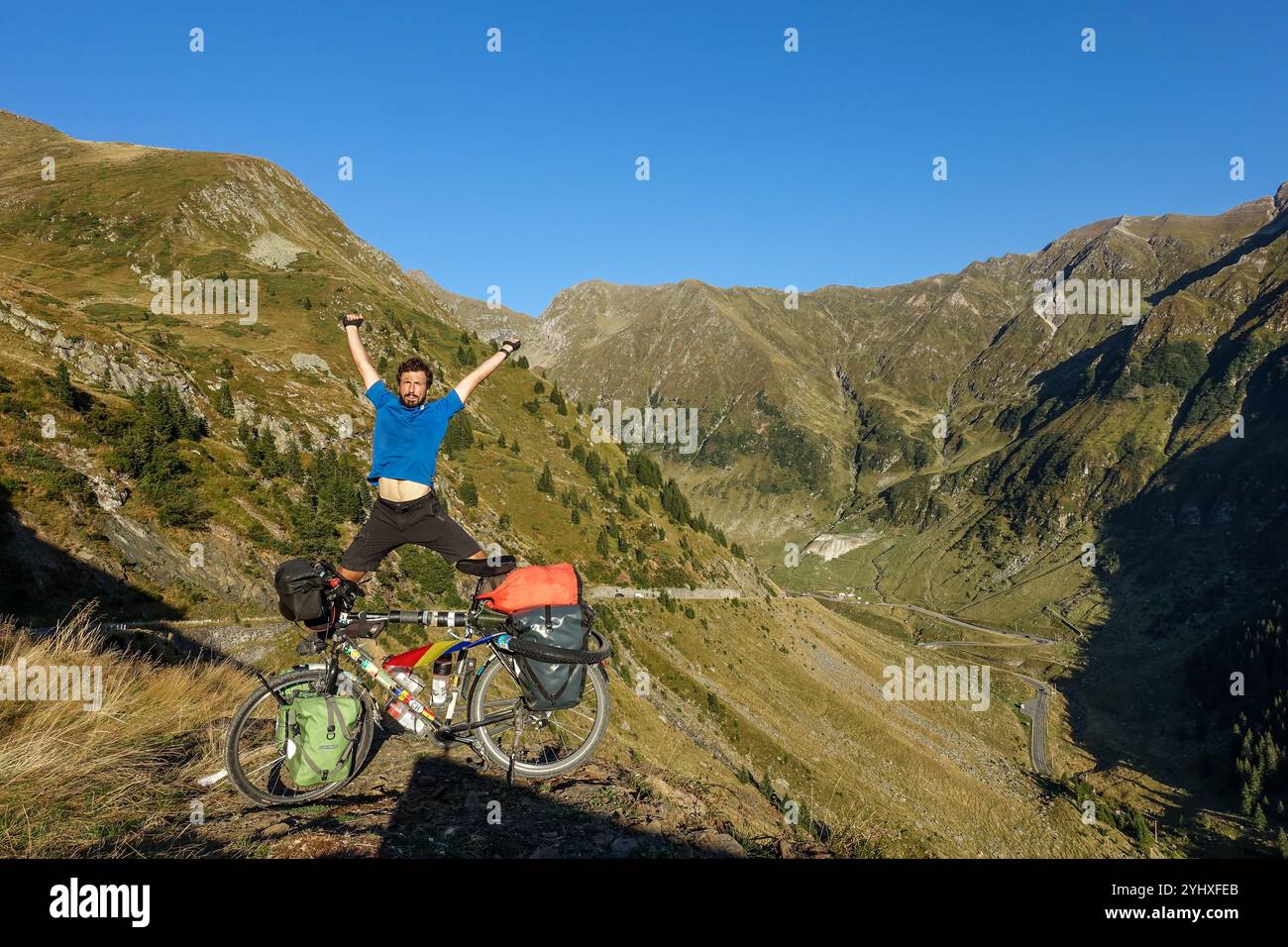 Un ciclista gioioso salta nell'emozione accanto a una bicicletta da turismo carica contro uno splendido paesaggio montano lungo la strada Transfagarasan, Romania Foto Stock