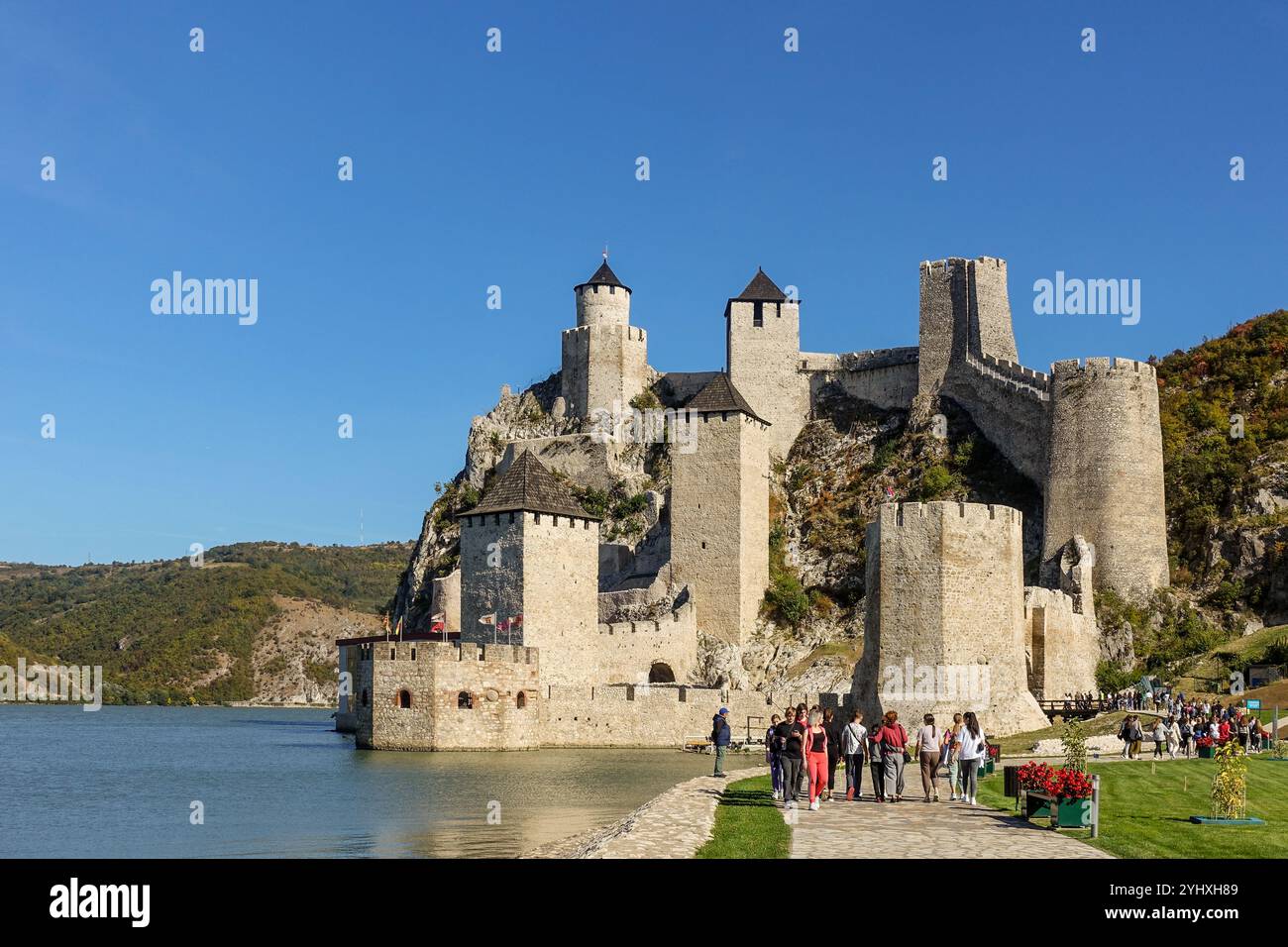 I turisti camminano verso la fortezza medievale di Golubac, con torri multiple su una collina rocciosa accanto a un fiume sotto un cielo azzurro Foto Stock