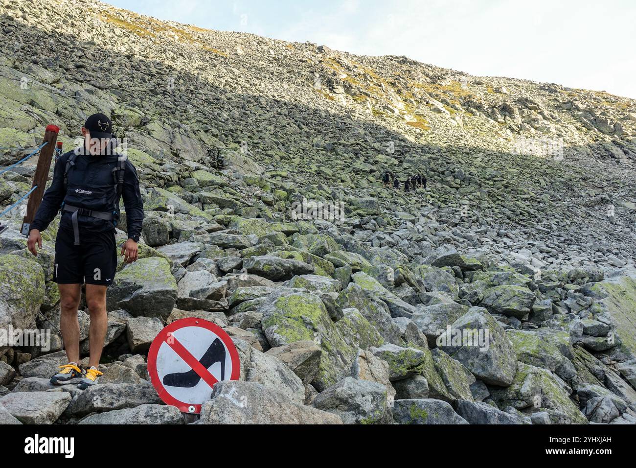 Un escursionista si erge in mezzo a un sentiero roccioso di montagna accanto a un cartello che scherzosamente proibisce scarpe con tacco alto, High Tatra, Slovacchia Foto Stock