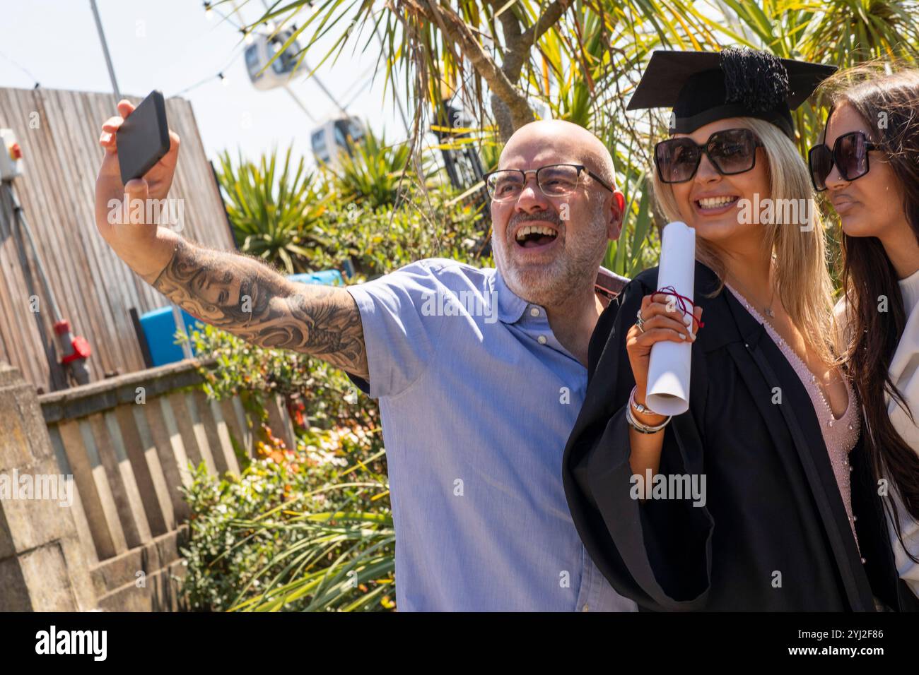 Un gioioso laureato con un berretto e un abito si fa un selfie con un padre e una sorella che si aggirano sotto un cielo soleggiato. Foto Stock