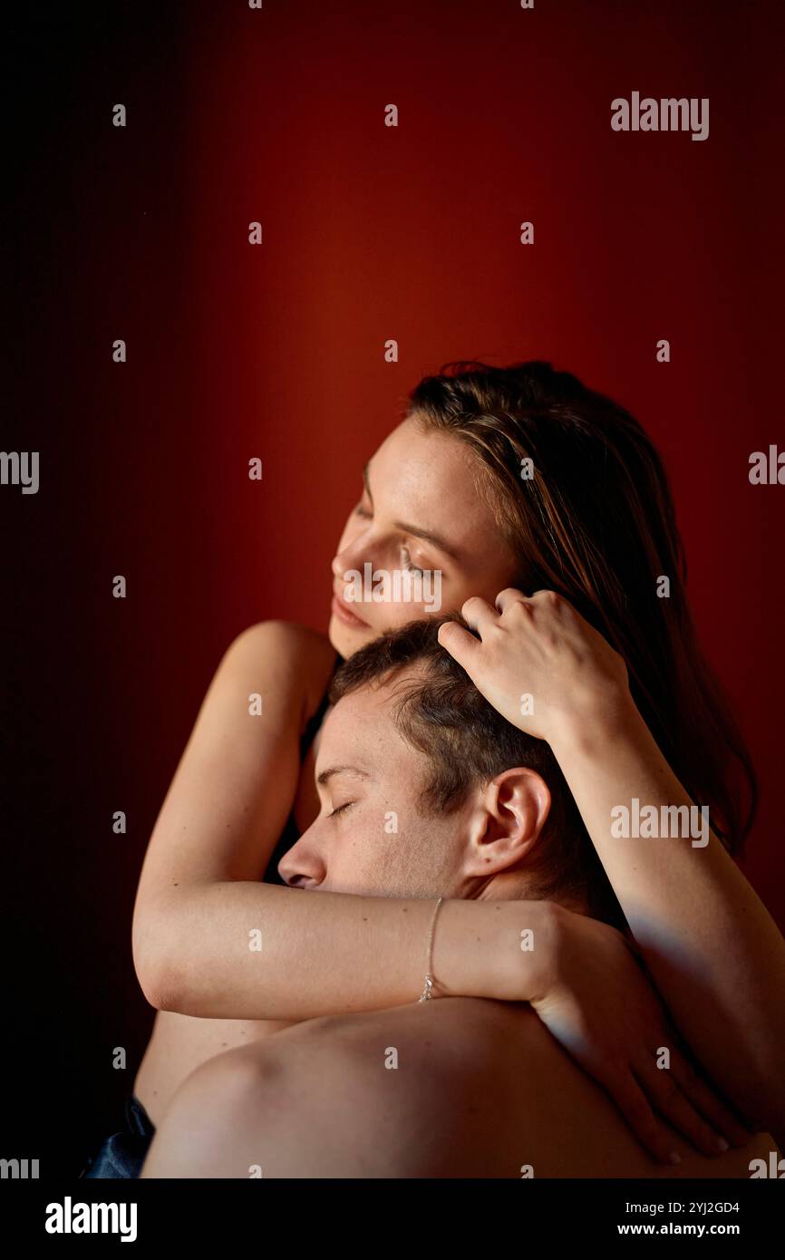 Una donna abbraccia con tenerezza un uomo da dietro, entrambi con espressioni introspettive su un caldo sfondo rosso. Foto Stock Una donna abbraccia con tenerezza un uomo da dietro, entrambi con espressioni introspettive su un caldo sfondo rosso. Foto Stock