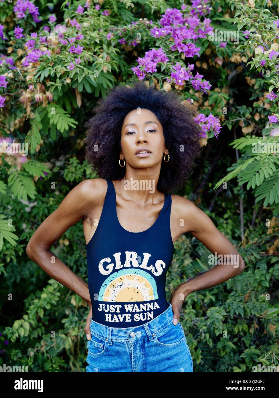 Donna sicura di sé con un afro sorprendente si erge davanti a un cespuglio di fiori viola, indossando una canotta e jeans blu. Foto Stock