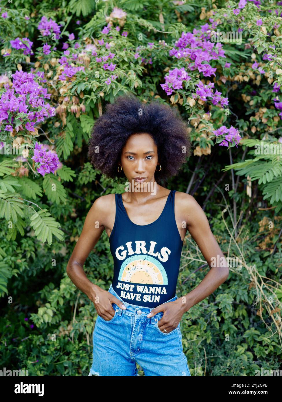 Donna sicura di sé con un afro sorprendente si erge davanti a un cespuglio di fiori viola, indossando una canotta e jeans blu. Foto Stock