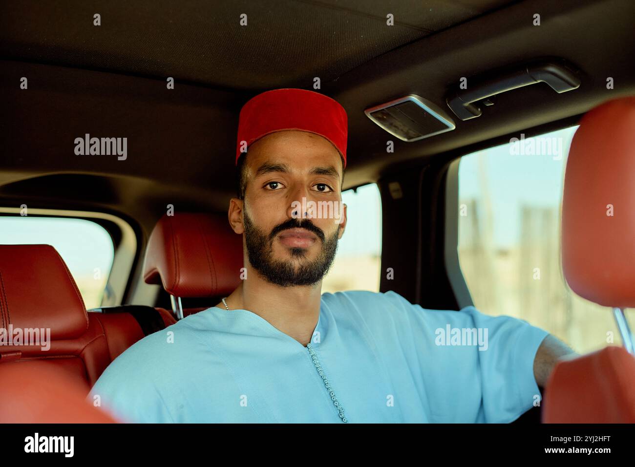 Giovane sicuro di sé con una camicia blu e un berretto rosso seduti in un'auto con un elegante interno in pelle rossa, Marocco Foto Stock