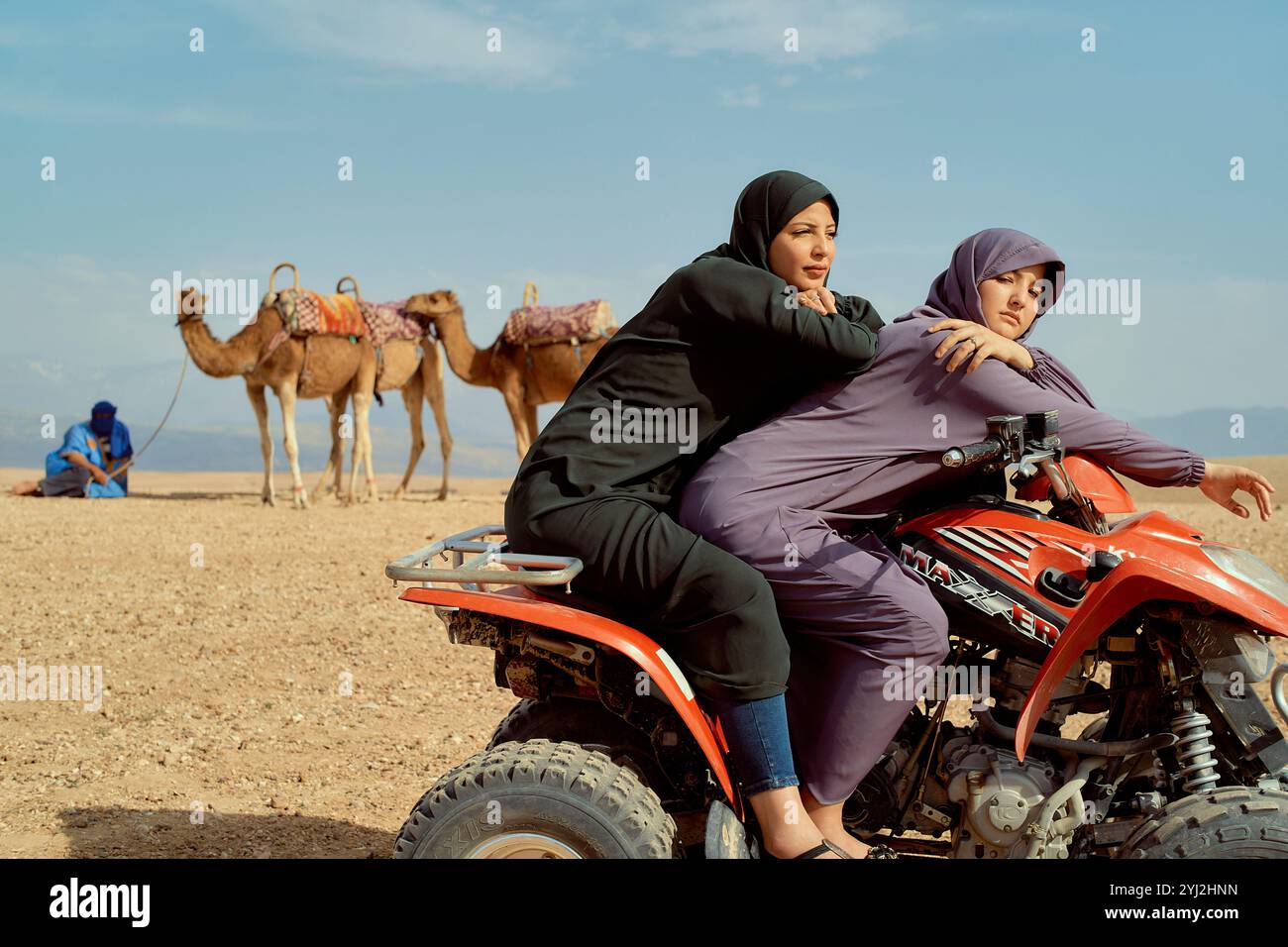 Due donne in hijab sedute su un ATV nel deserto con cammelli e una persona sullo sfondo, il Marocco Foto Stock Due donne in hijab sedute su un ATV nel deserto con cammelli e una persona sullo sfondo, il Marocco Foto Stock