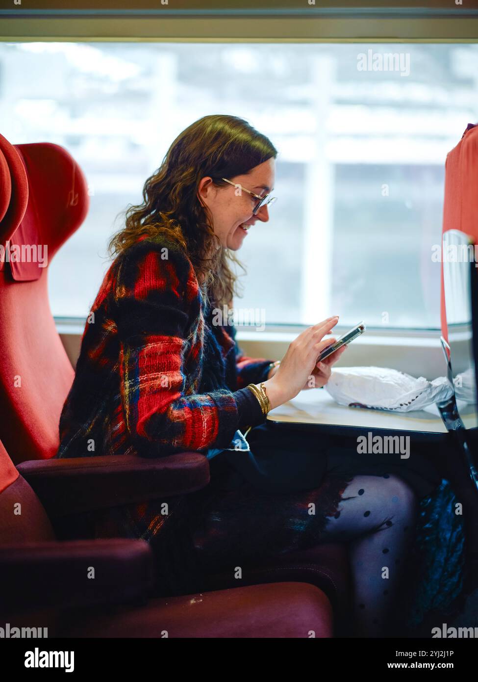 Donna sorridente che utilizza uno smartphone seduto in uno scomparto treno con sedili rossi. Foto Stock