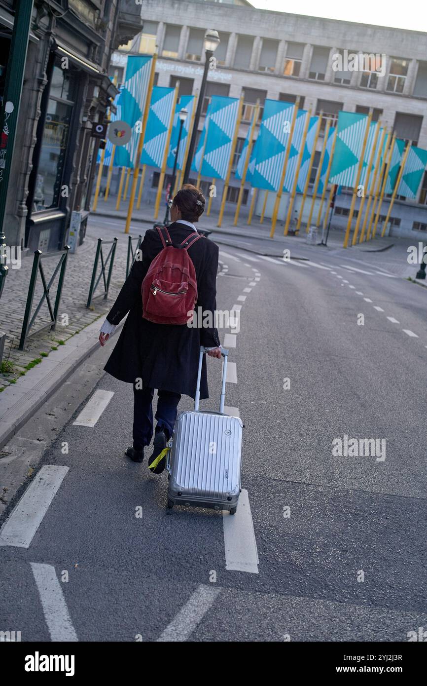 Donna che cammina lungo una strada tirando una valigia bianca con uno zaino rosso in una zona urbana Foto Stock