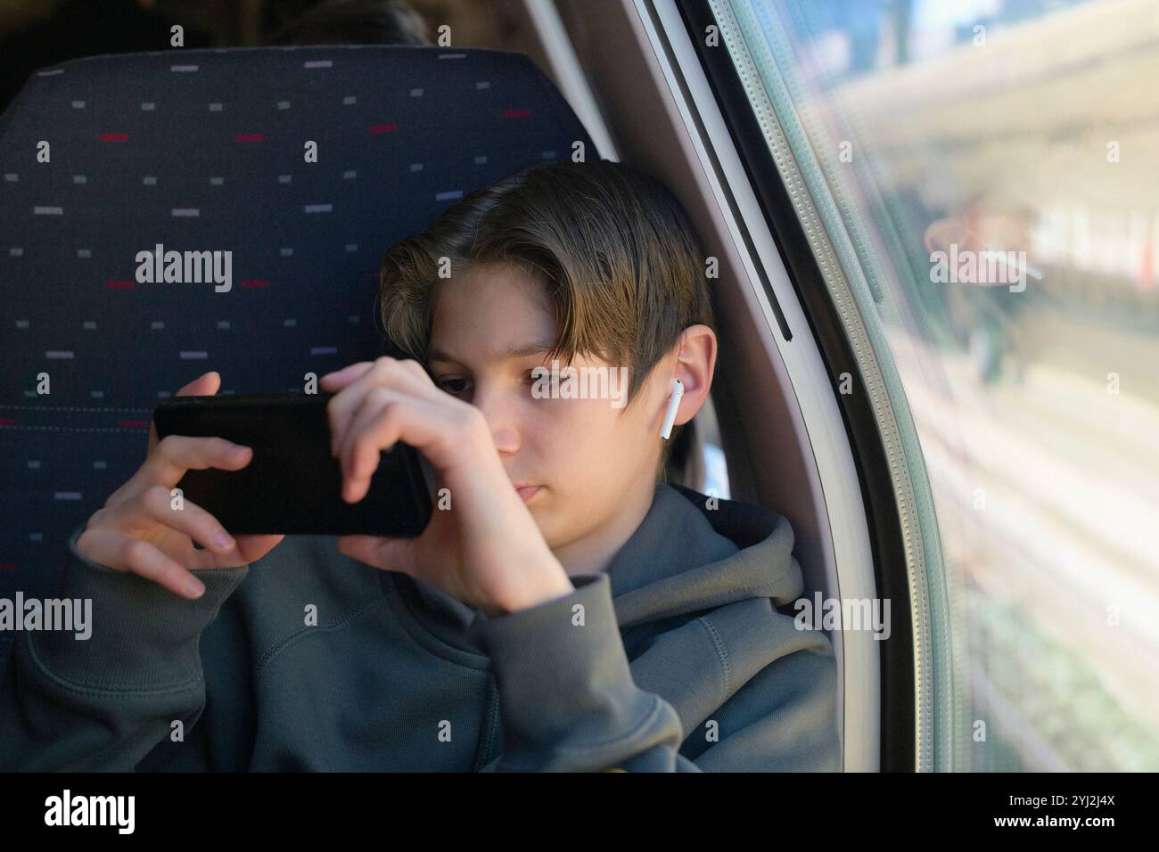 Un adolescente che indossa gli auricolari wireless si concentra con attenzione sul suo smartphone mentre è seduto accanto alla finestra su un treno. Foto Stock