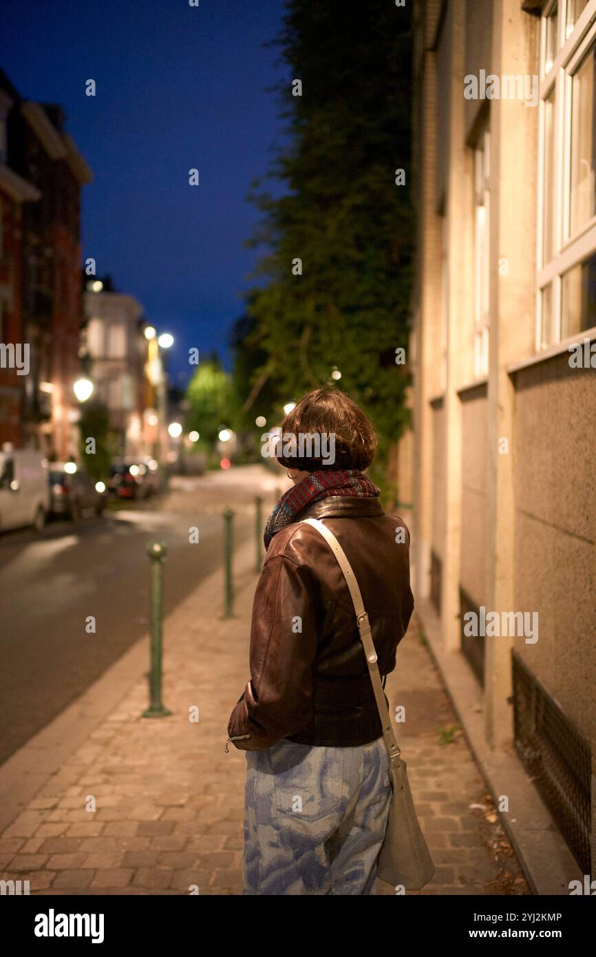 Una giovane donna con giacca e sciarpa in pelle si trova in una strada di città di notte, rivolta lontano dalla fotocamera. Foto Stock