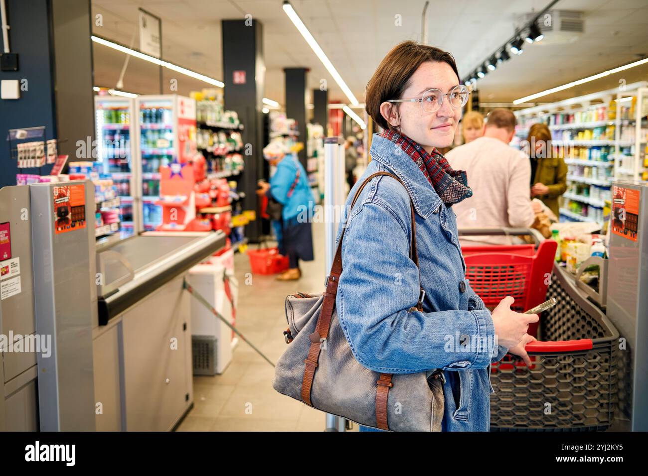 Donna con occhiali e giacca in denim che spinge un carrello nella corsia di un negozio di alimentari. Foto Stock