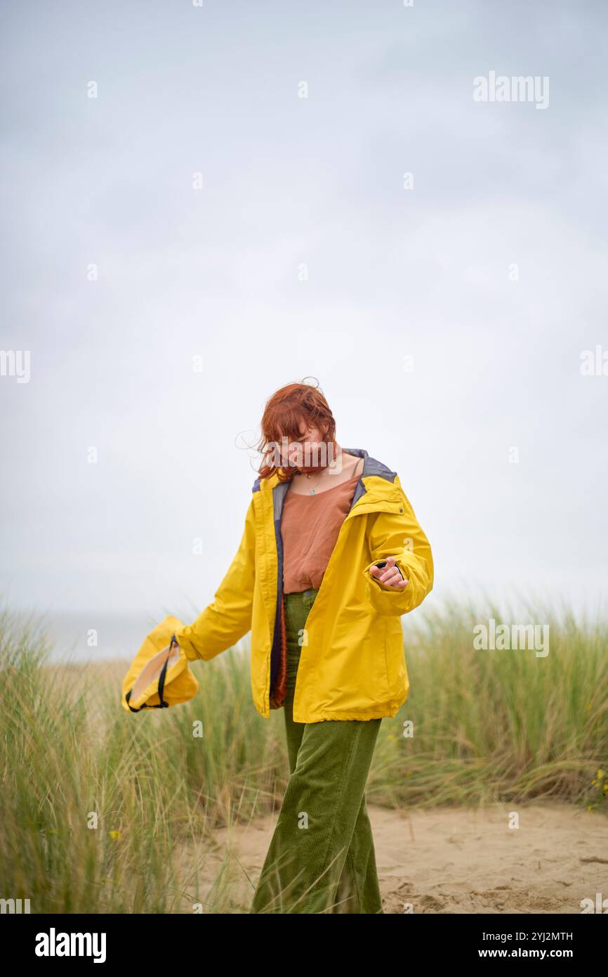 Una persona che indossa un impermeabile giallo si erge su una spiaggia sabbiosa, guardando in basso con i capelli spazzati dal vento, Belgio Foto Stock