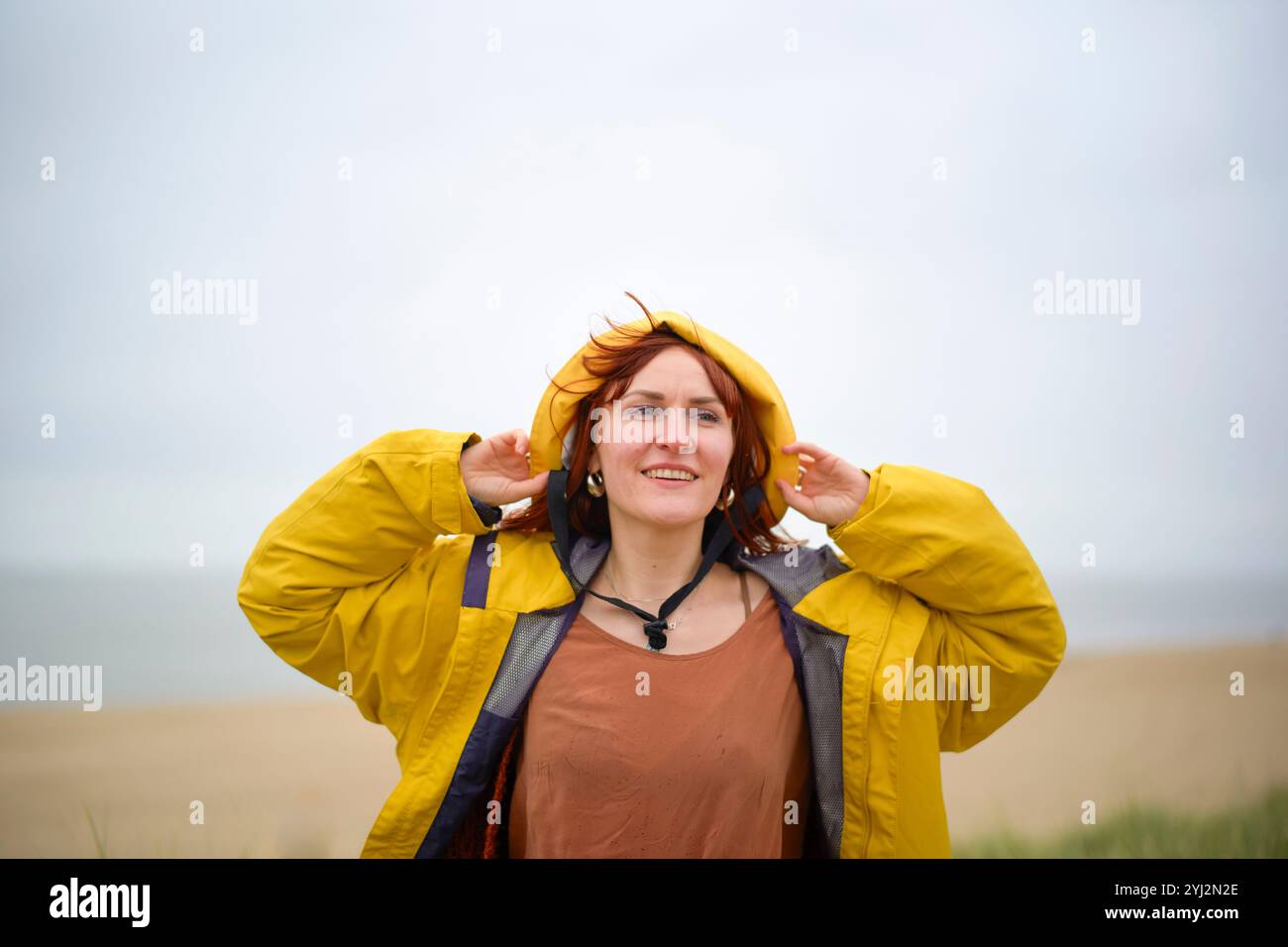 Donna sorridente in un impermeabile giallo in piedi su una spiaggia con cieli nuvolosi, Belgio Foto Stock