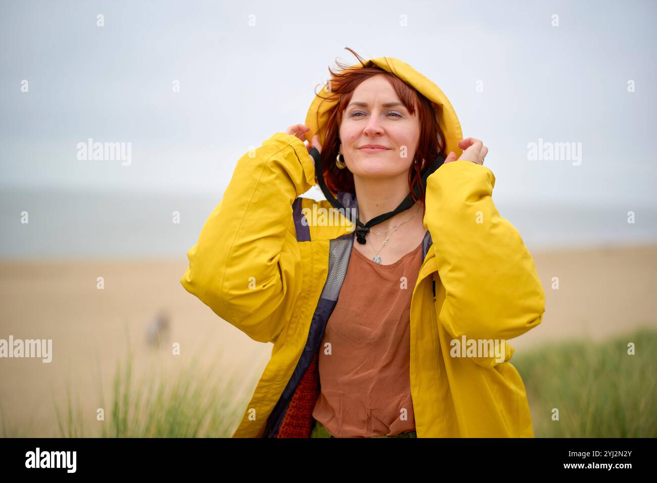 Donna sorridente in un impermeabile giallo in piedi su una spiaggia con dune sullo sfondo, Belgio Foto Stock