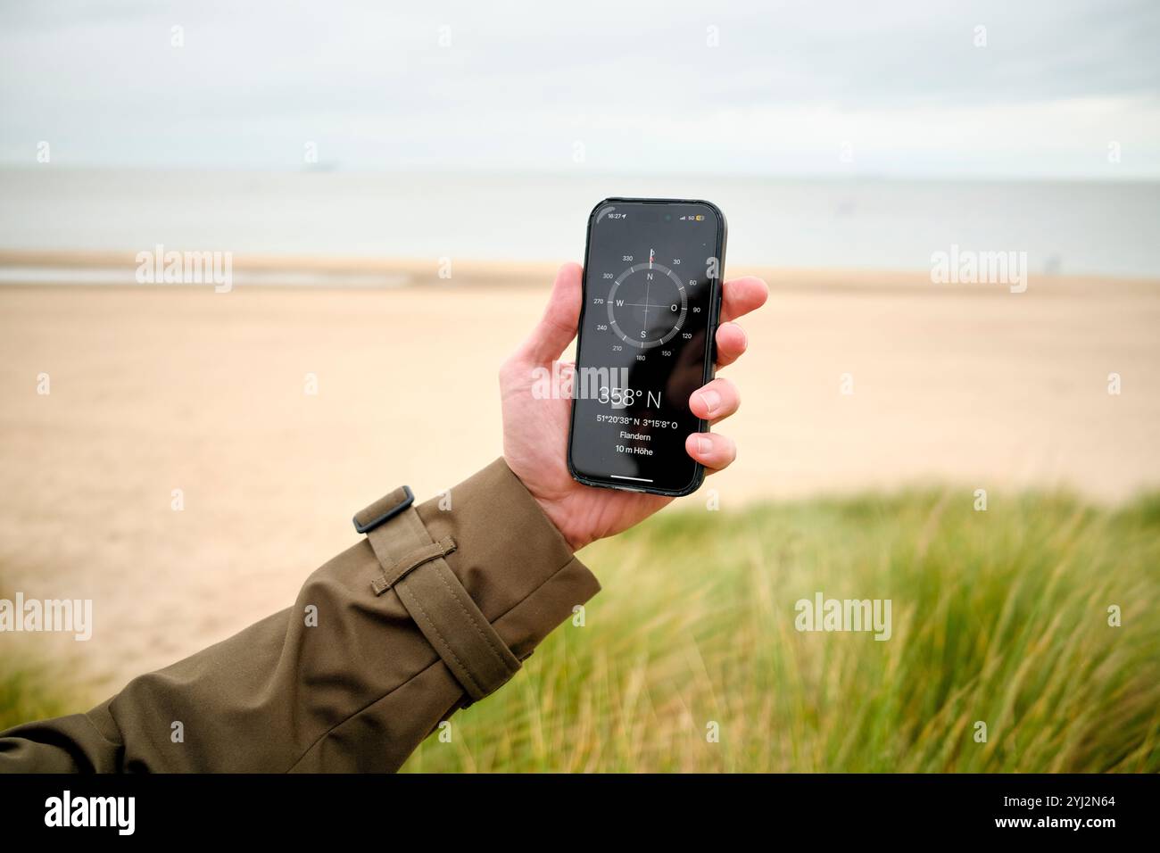 Mano che tiene in mano uno smartphone con un'app bussola sullo schermo di una spiaggia sabbiosa sotto un cielo nuvoloso, Belgio Foto Stock