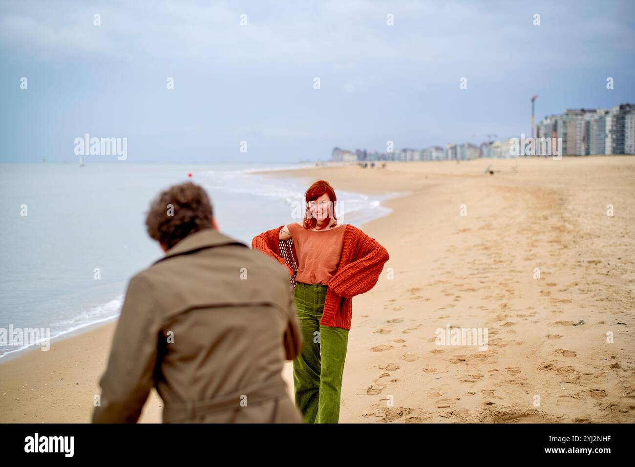 Una donna con i capelli rossi sorride alla telecamera su una spiaggia sabbiosa con una figura maschile sfocata che passa davanti e edifici sullo sfondo, il Belgio Foto Stock