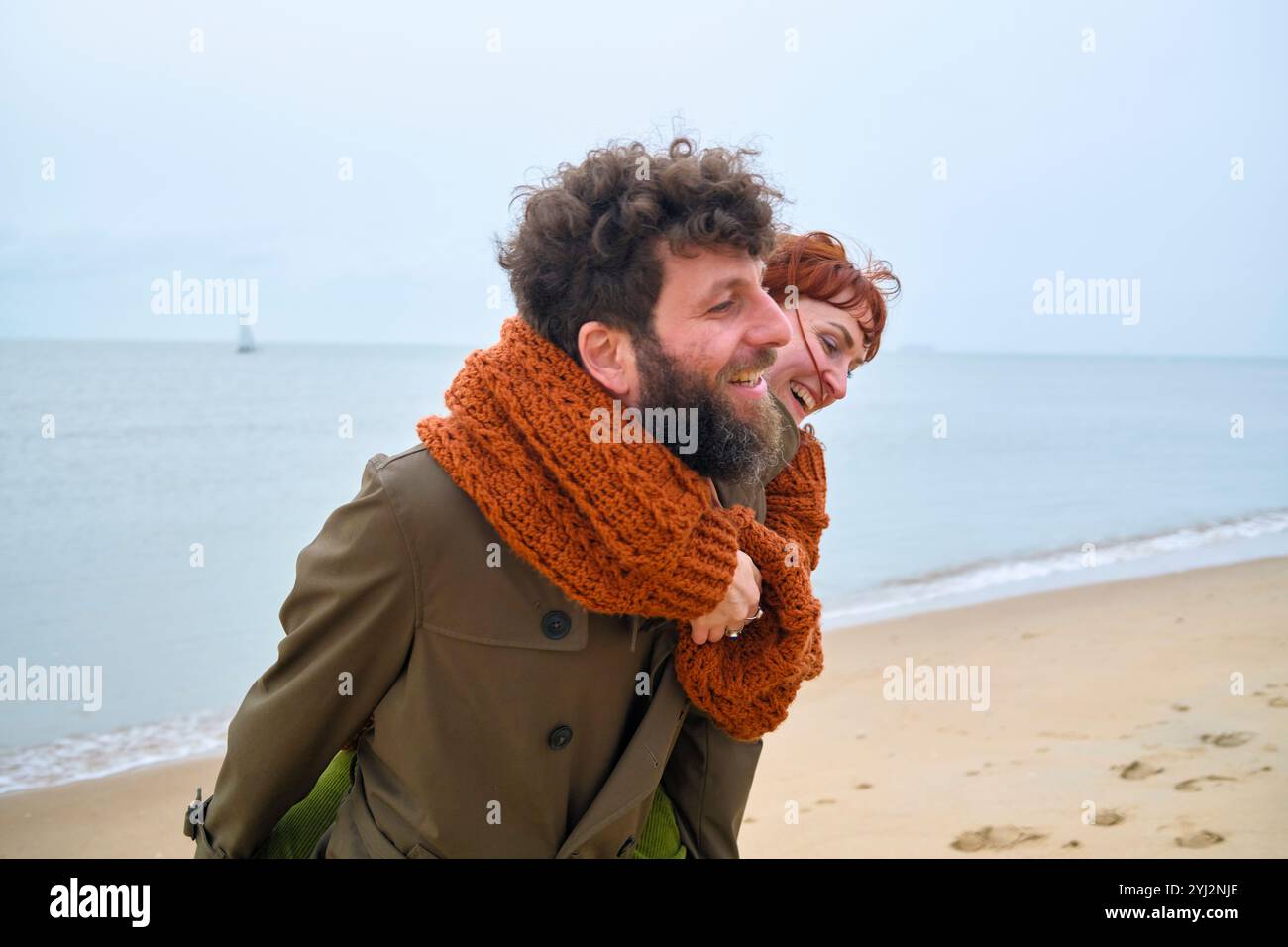 Uomo sorridente con la barba che fa un giro in piggyback a una donna felice in una giornata nuvolosa sulla spiaggia, in Belgio Foto Stock