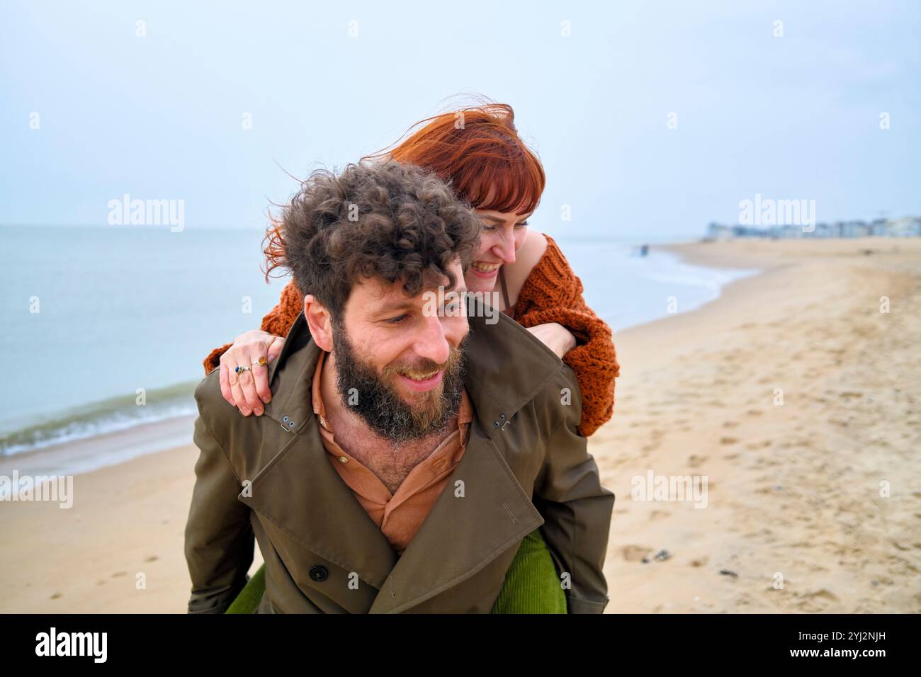 Uomo sorridente con la barba che fa un giro in piggyback a una donna felice in una giornata nuvolosa sulla spiaggia, in Belgio Foto Stock