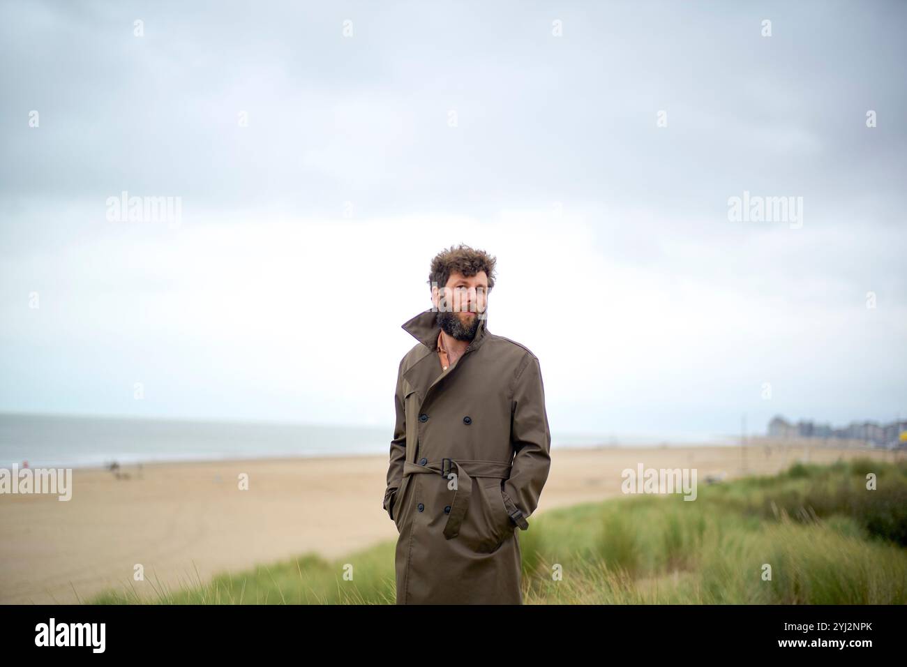 Uomo in trench coat in piedi su dune di sabbia con cieli nuvolosi e spiaggia sullo sfondo, Belgio Foto Stock