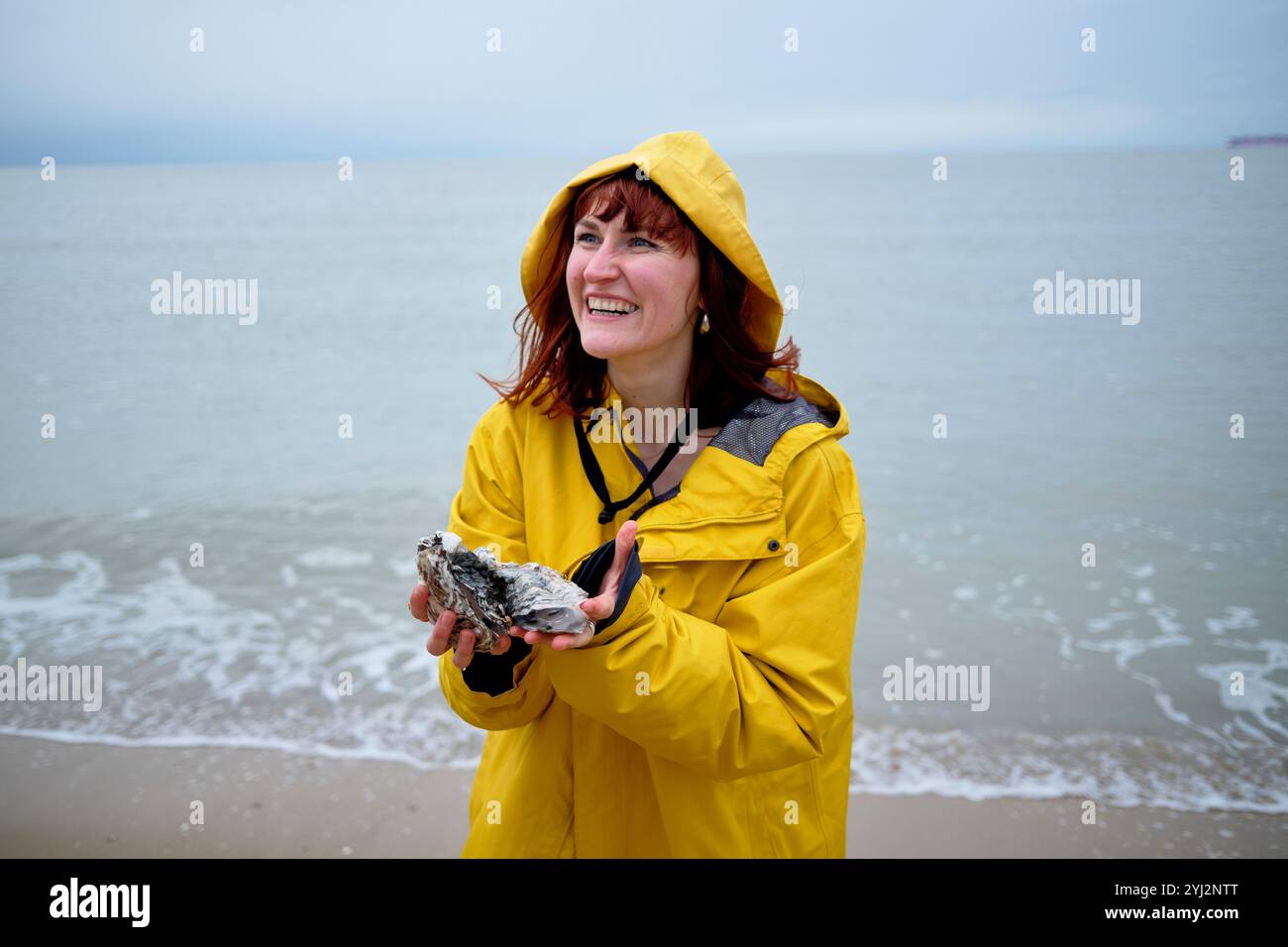 Donna sorridente in impermeabile giallo che tiene in mano un'ostrica in spiaggia in un giorno coperto, in Belgio Foto Stock