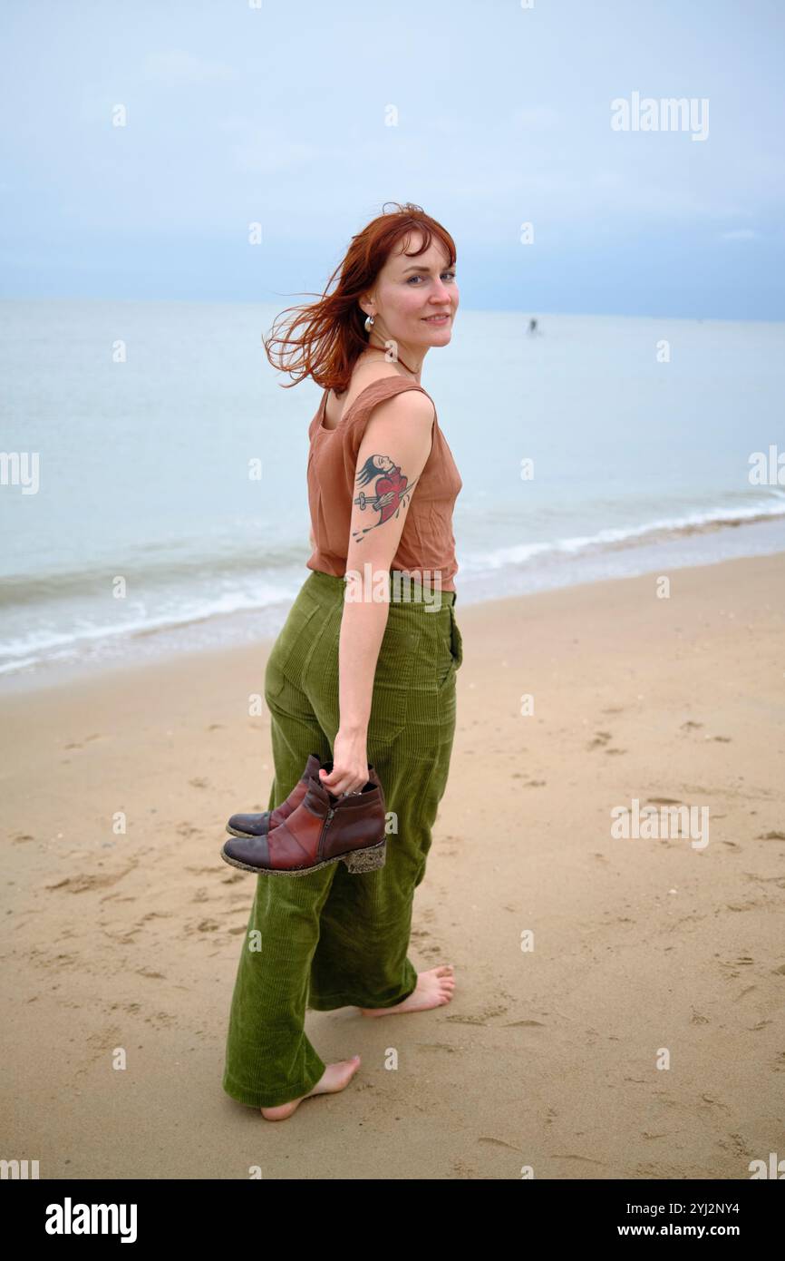 Donna con i capelli rossi e un tatuaggio sul braccio cammina a piedi nudi su una spiaggia sabbiosa, tenendo le scarpe in una mano e guardando le spalle, Belgio Foto Stock